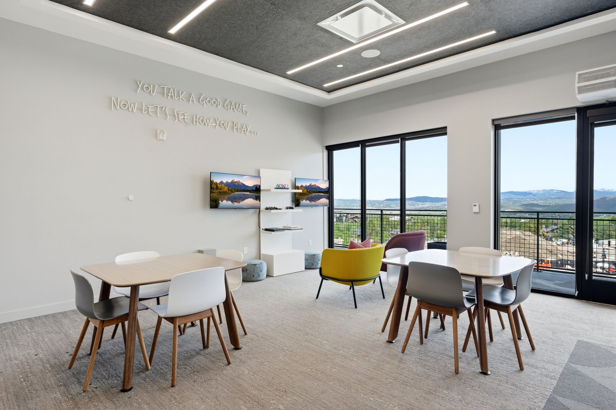 Open-concept common area featuring expansive full-height windows, wall-mounted display screens, contemporary tables, and built-in linear ceiling lighting