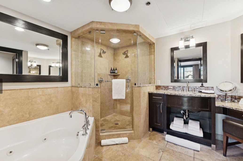 Spacious bathroom featuring a jetted soaking tub, a glass-enclosed shower with dual showerheads, stone tile flooring, and a vanity with a granite countertop