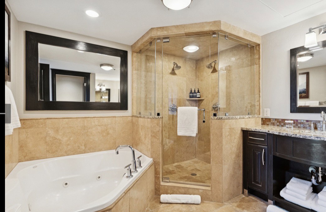 Spacious bathroom featuring a corner jetted soaking tub, a multi-head glass-enclosed shower, a vanity with granite countertop, and stone-look tile flooring and surrounds