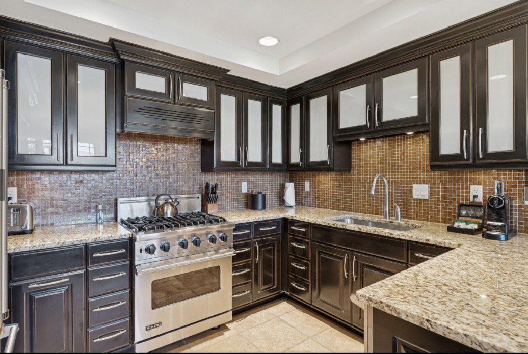 Gourmet kitchen featuring dark wood cabinetry with frosted glass inserts, speckled light-toned countertops, a mosaic tile backsplash, stainless steel gas range, and a stainless steel sink with a gooseneck faucet