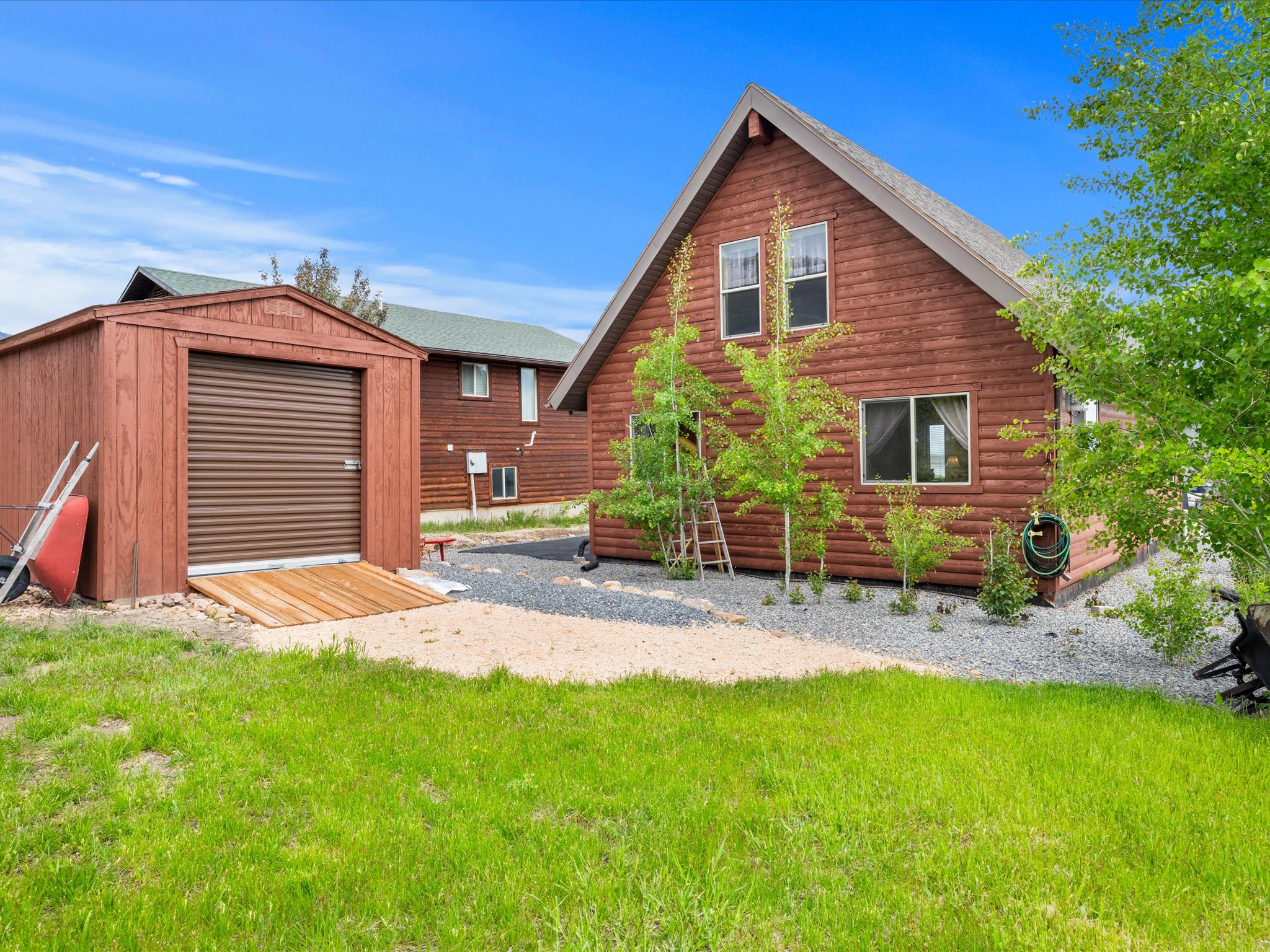 Wood-sided storage shed with a roll-up door and ramp, accompanied by a main residence featuring log siding and a gravel perimeter