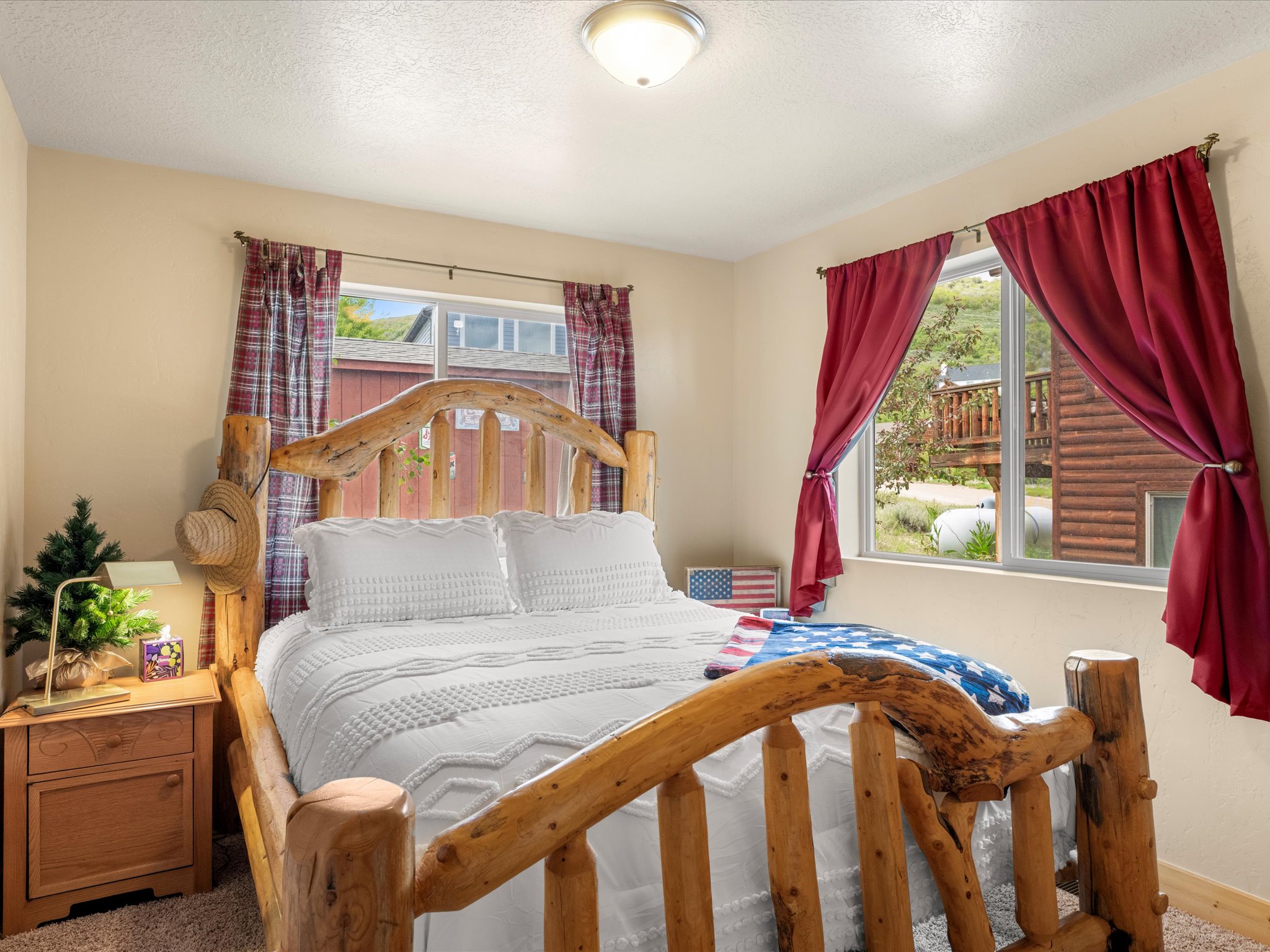 Bedroom featuring a natural wood bed frame, a window with red drapery, a wood-finish nightstand, and a flush-mount ceiling light fixture