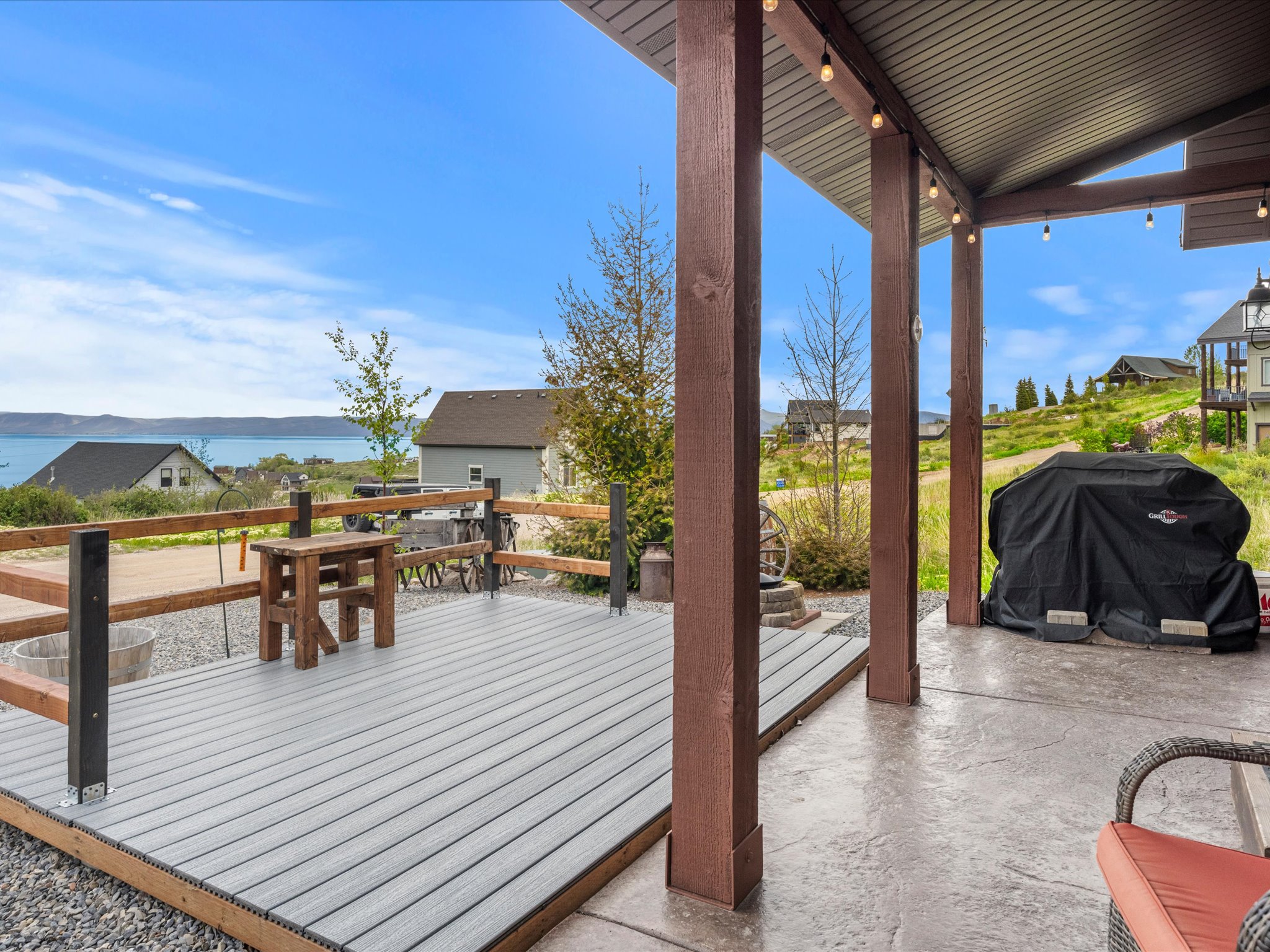 Spacious outdoor living area featuring a covered patio with string lighting, a composite deck with wood railing, and a stamped concrete patio, all overlooking a prominent body of water