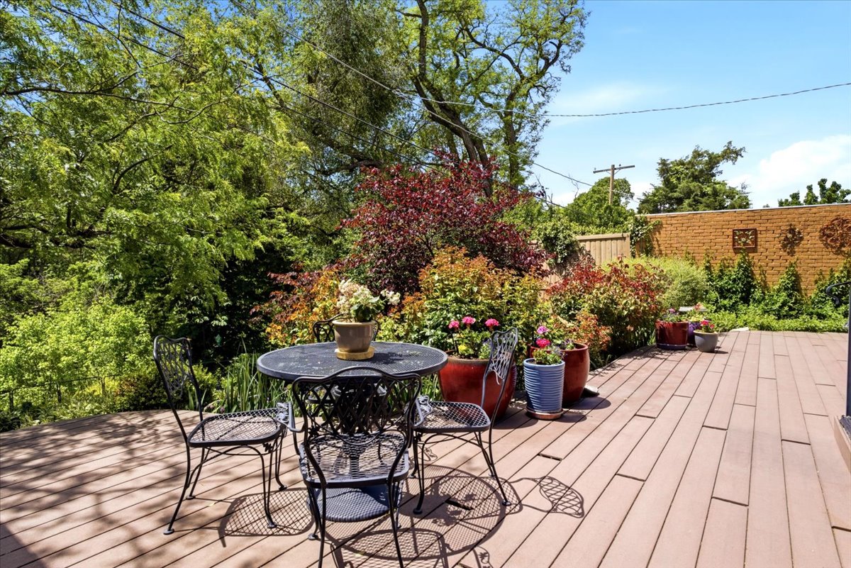 Expansive outdoor deck featuring wood-finish flooring, mature landscaping, varied foliage, and a decorative brick wall
