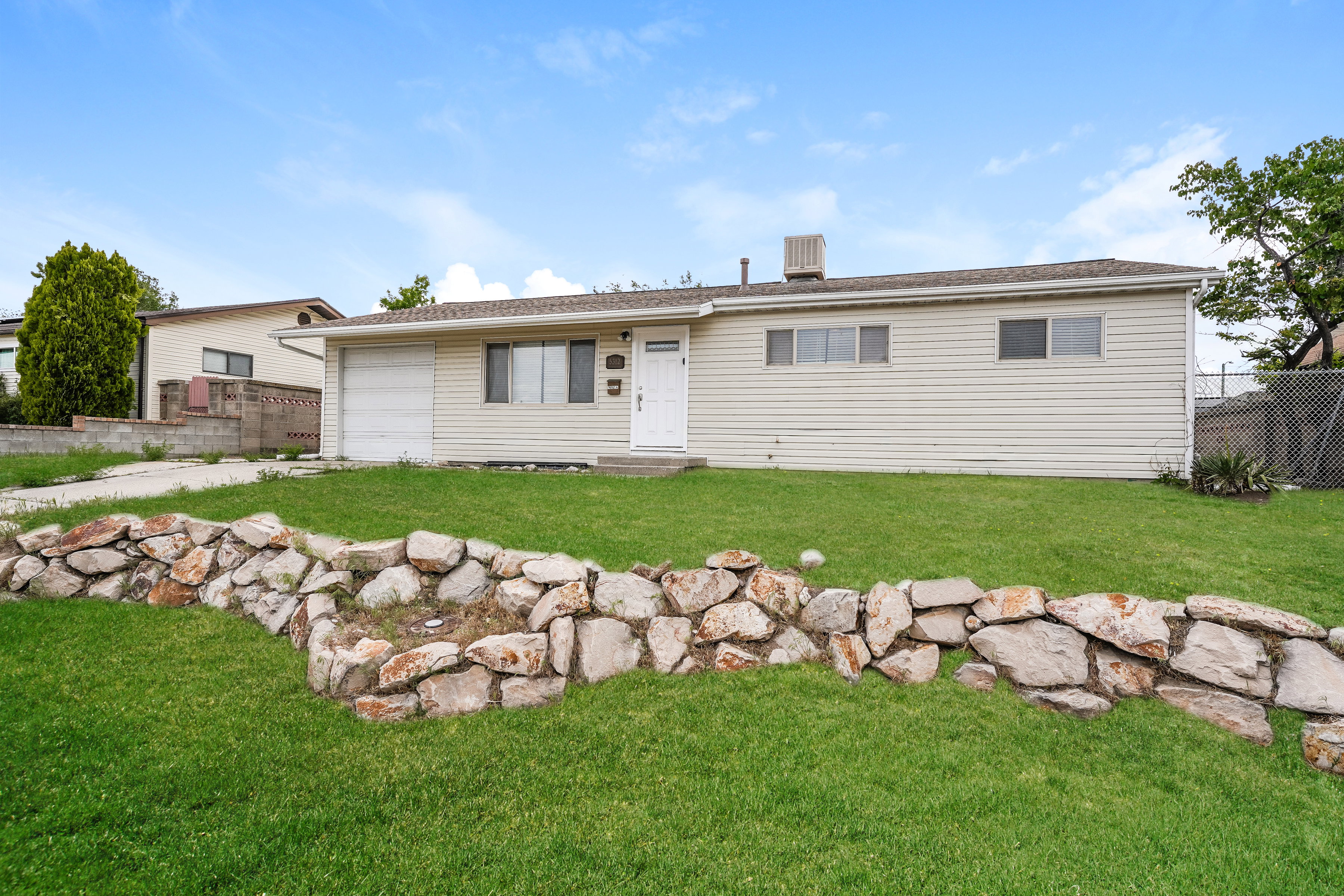 Ranch-style residence featuring a white facade, an attached garage, a white entry door, multiple windows, and a landscaped front yard with a rock retaining wall