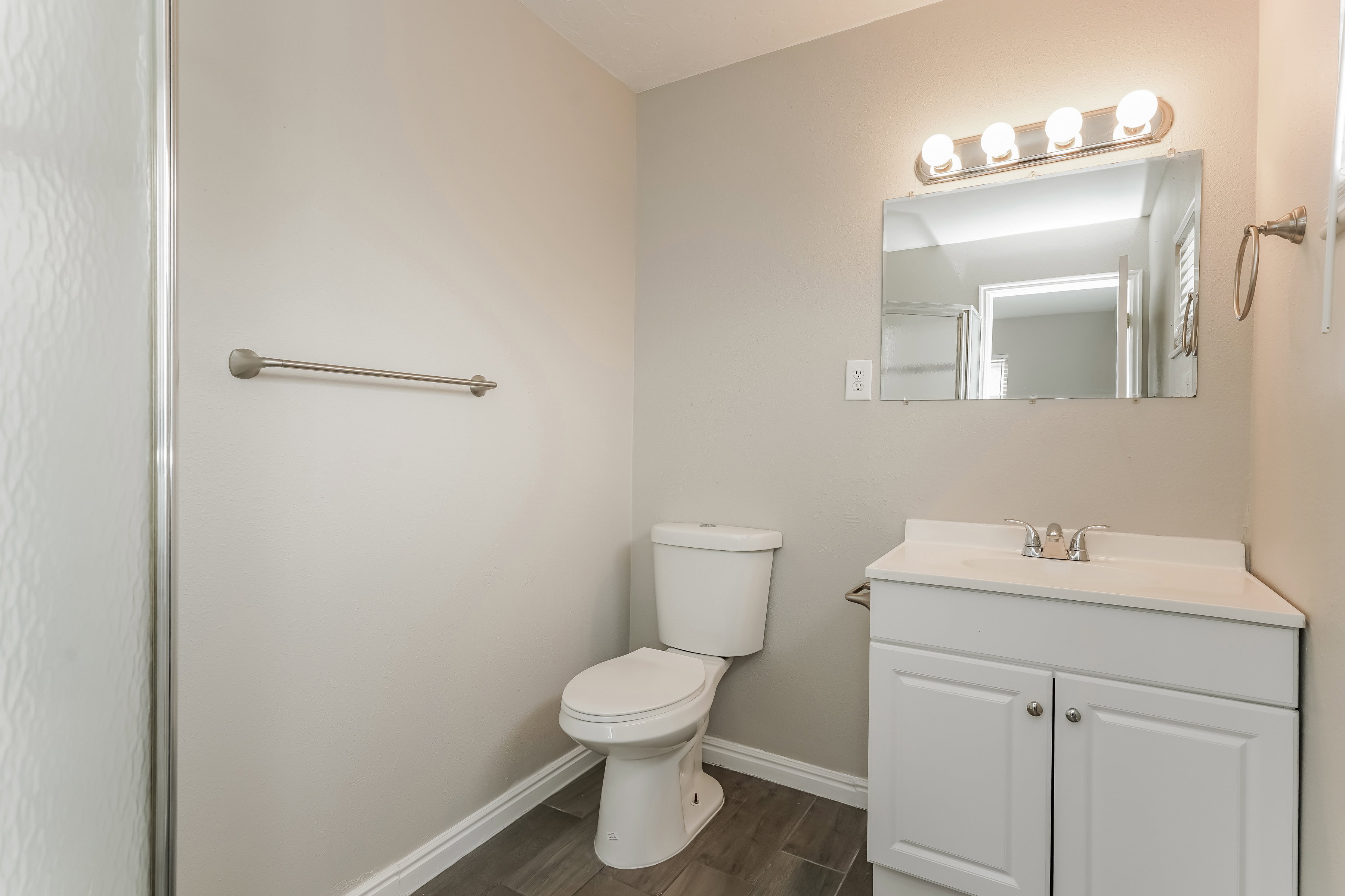 Bathroom featuring a single vanity with storage, a rectangular mirror with overhead lighting, a toilet, and wood-finish flooring