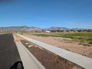 Expansive undeveloped lot featuring a paved road, concrete sidewalk, distant mountain range, and clear skies