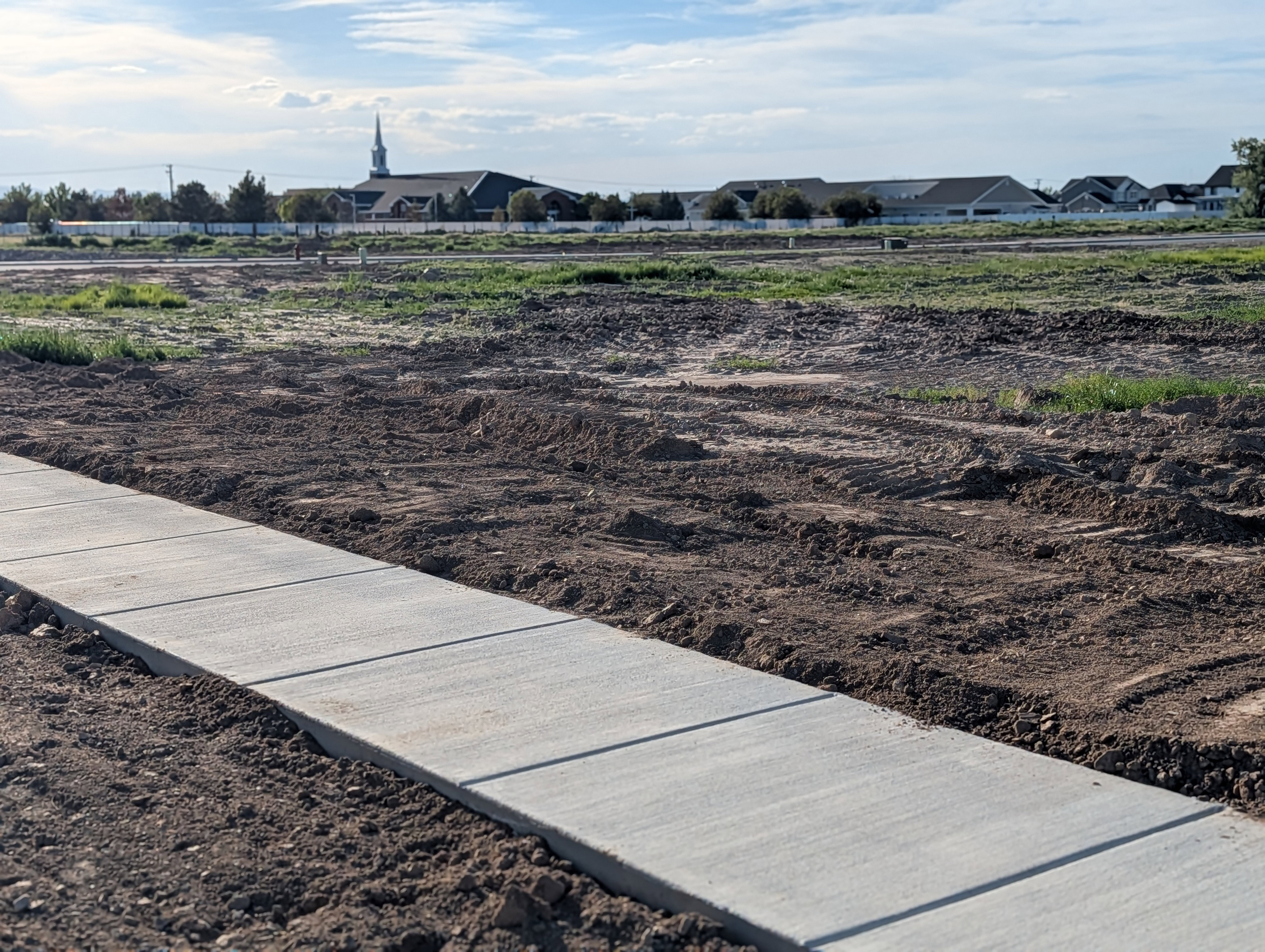 Concrete sidewalk bordering a graded parcel with distant structures visible under a partly cloudy sky