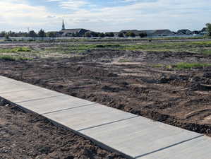 Concrete sidewalk bordering a graded parcel with distant structures visible under a partly cloudy sky