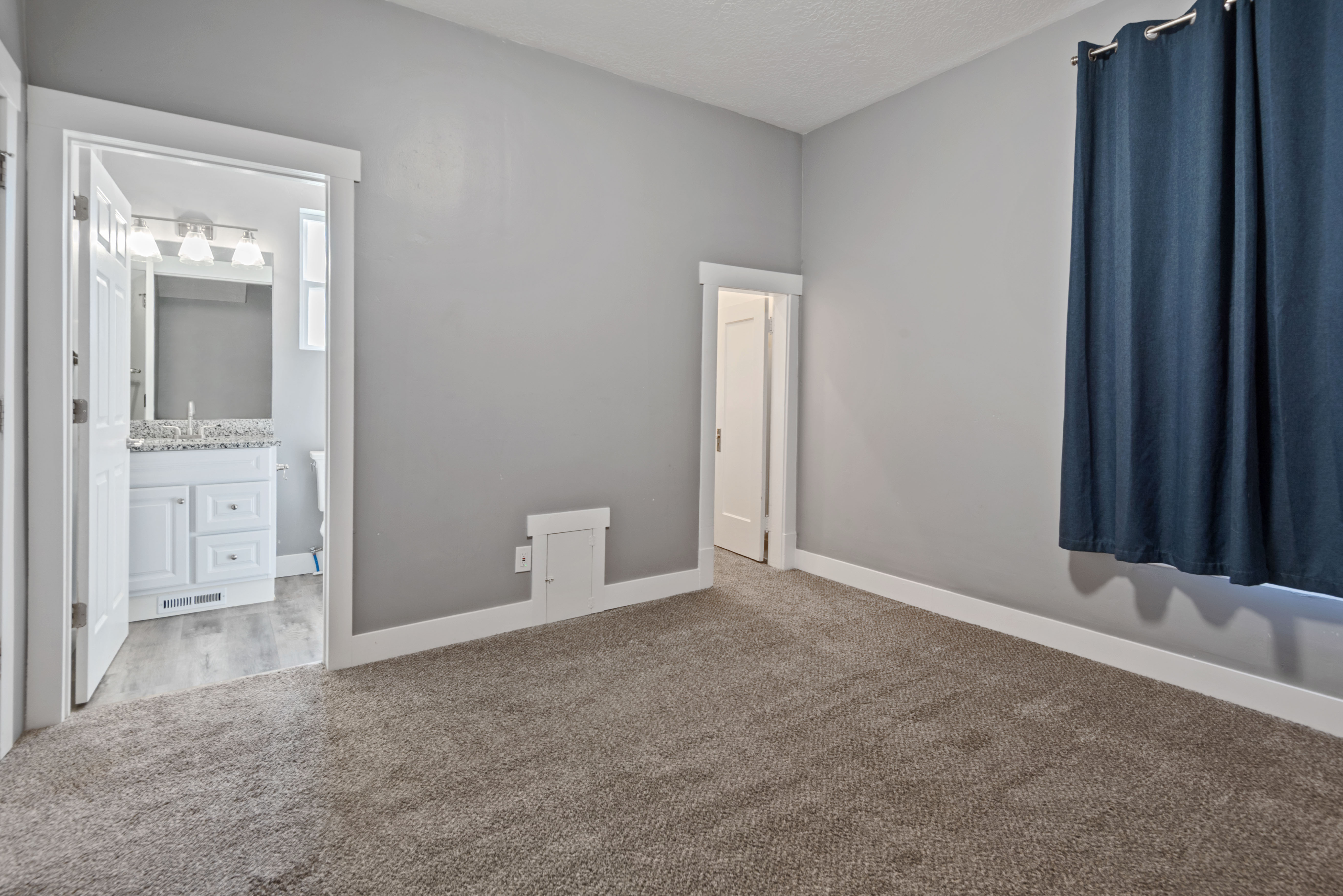Spacious room featuring neutral gray walls, plush carpet flooring, white baseboards, and a doorway opening to a bathroom