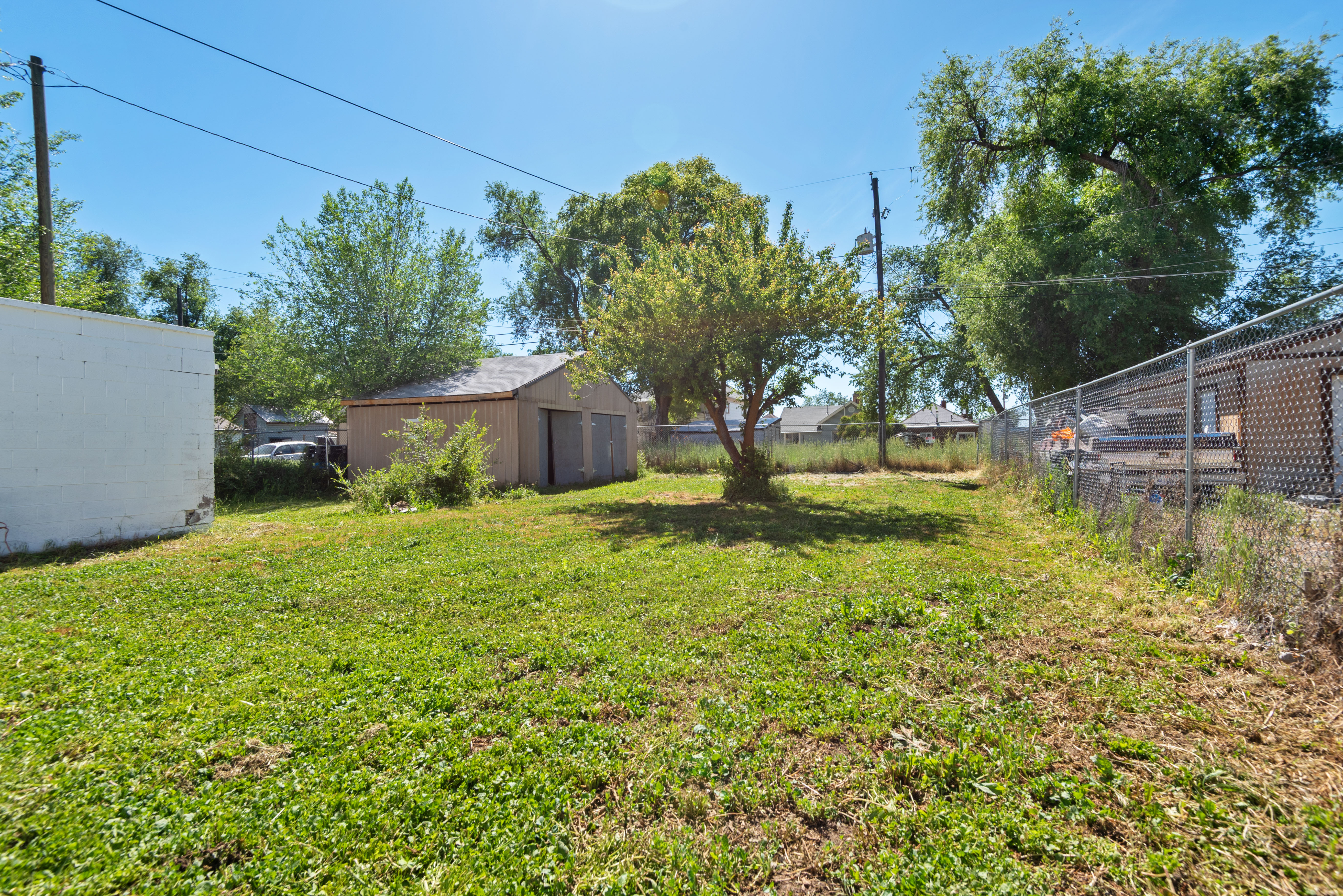 Expansive rear yard featuring green lawn, mature trees, a detached storage building with two garage doors, and perimeter chain link fencing