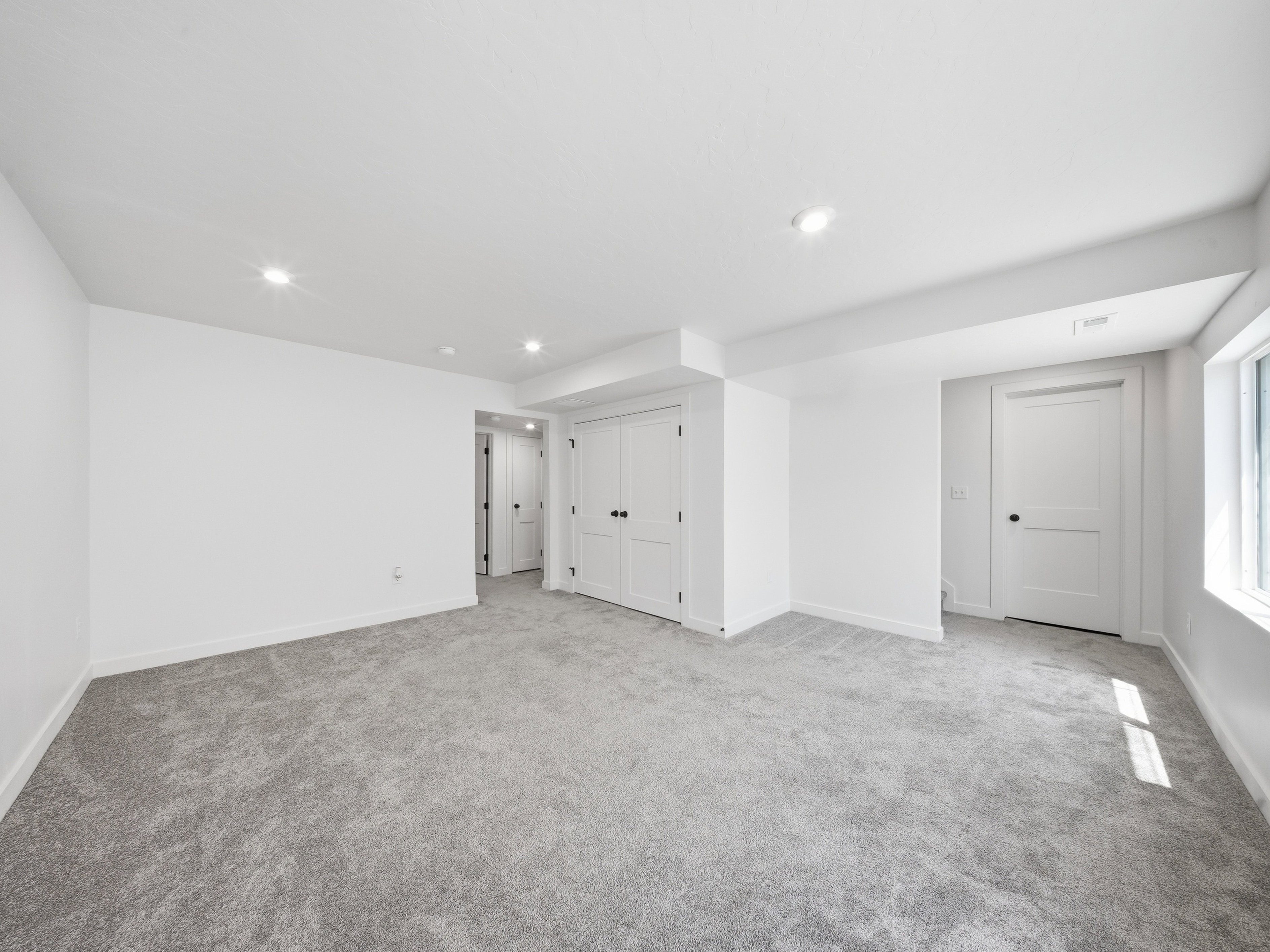 Spacious room featuring light grey carpet, white walls, recessed ceiling lights, and multiple white panel doors