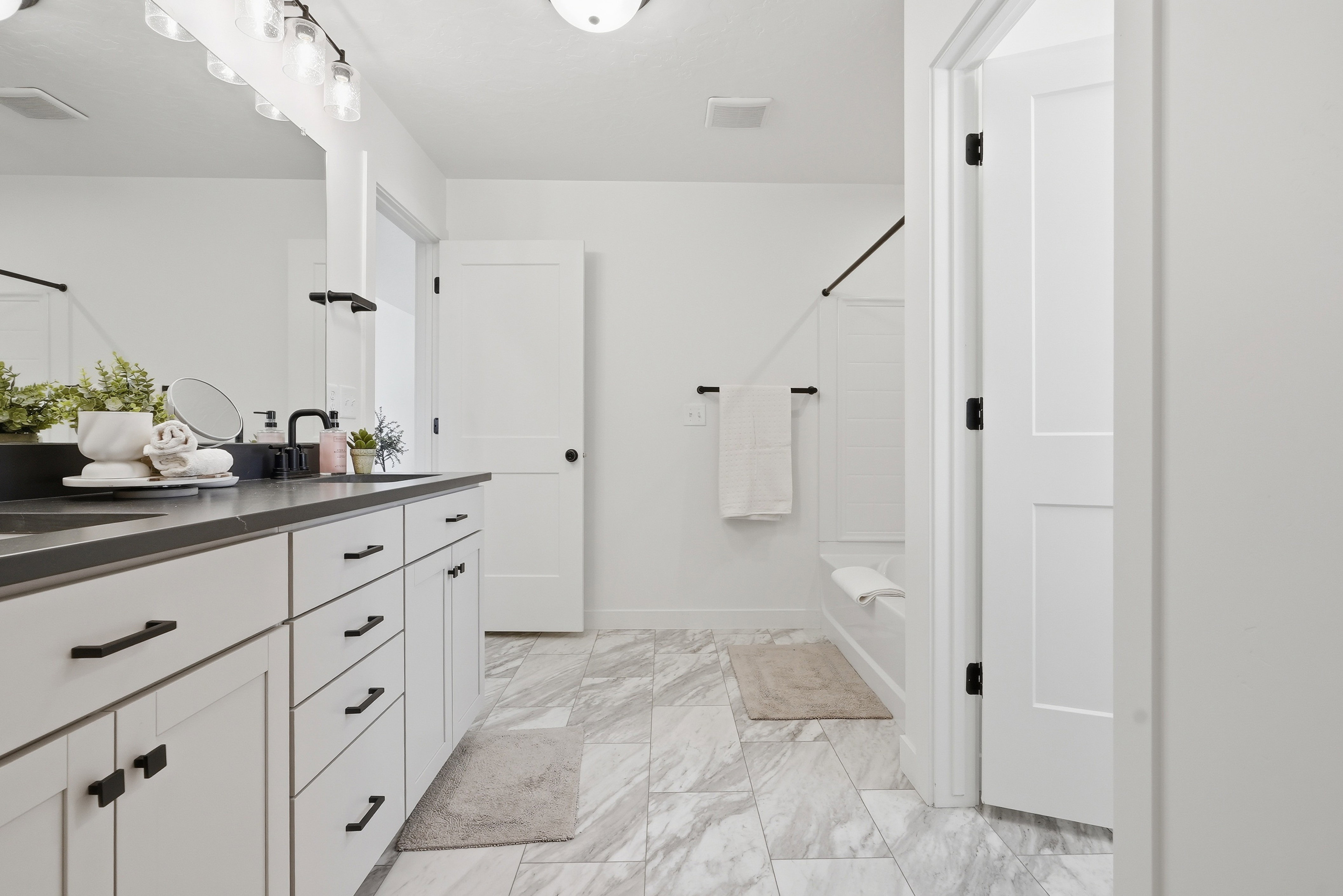 Bathroom featuring a dual vanity with a dark countertop and white cabinetry, marble-look floor tiling, a built-in bathtub with subway tile surround, and matte black fixtures