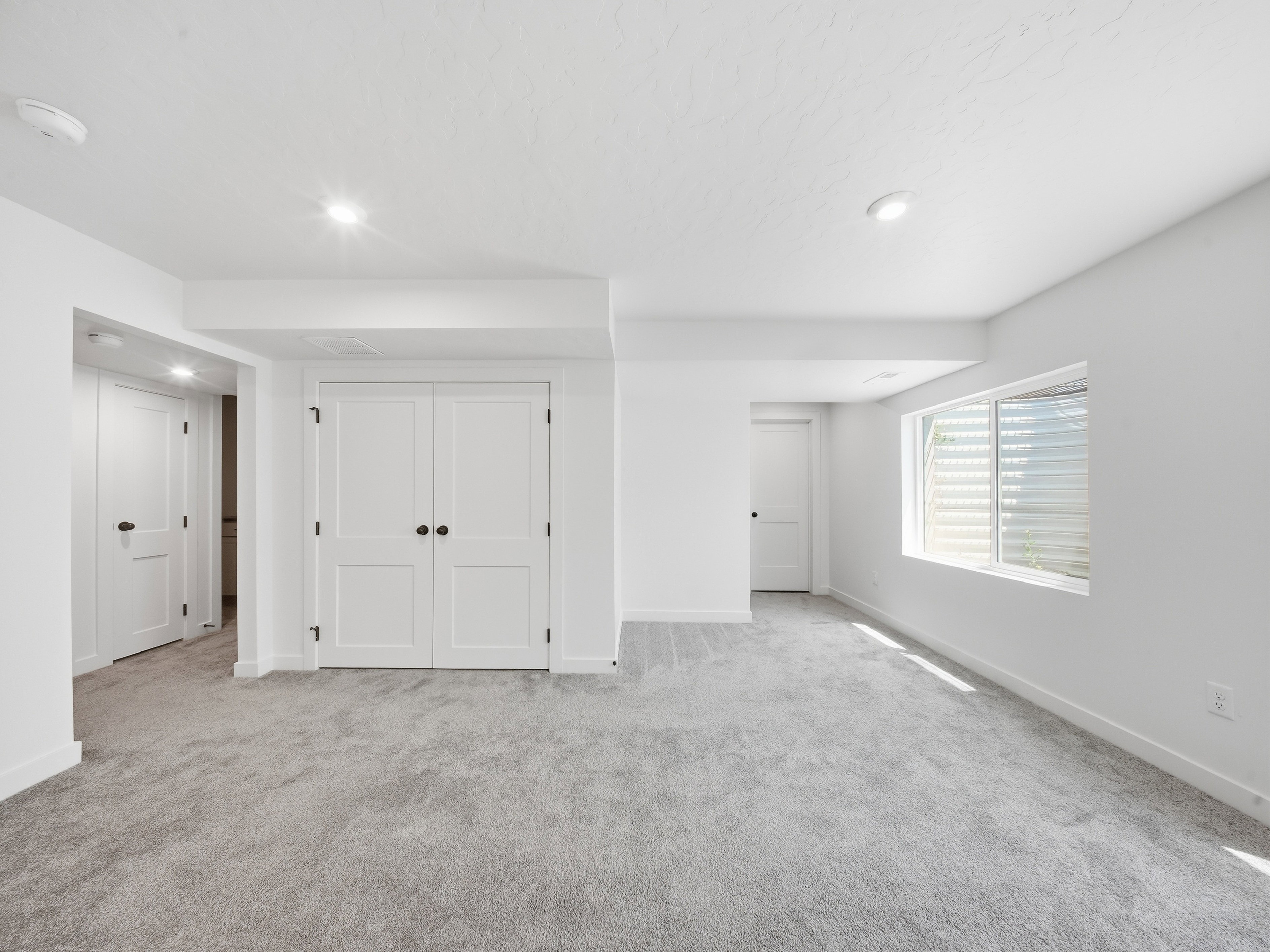 Spacious room featuring light gray carpeting, recessed ceiling lighting, a window with horizontal blinds, and white paneled doors with matte black hardware