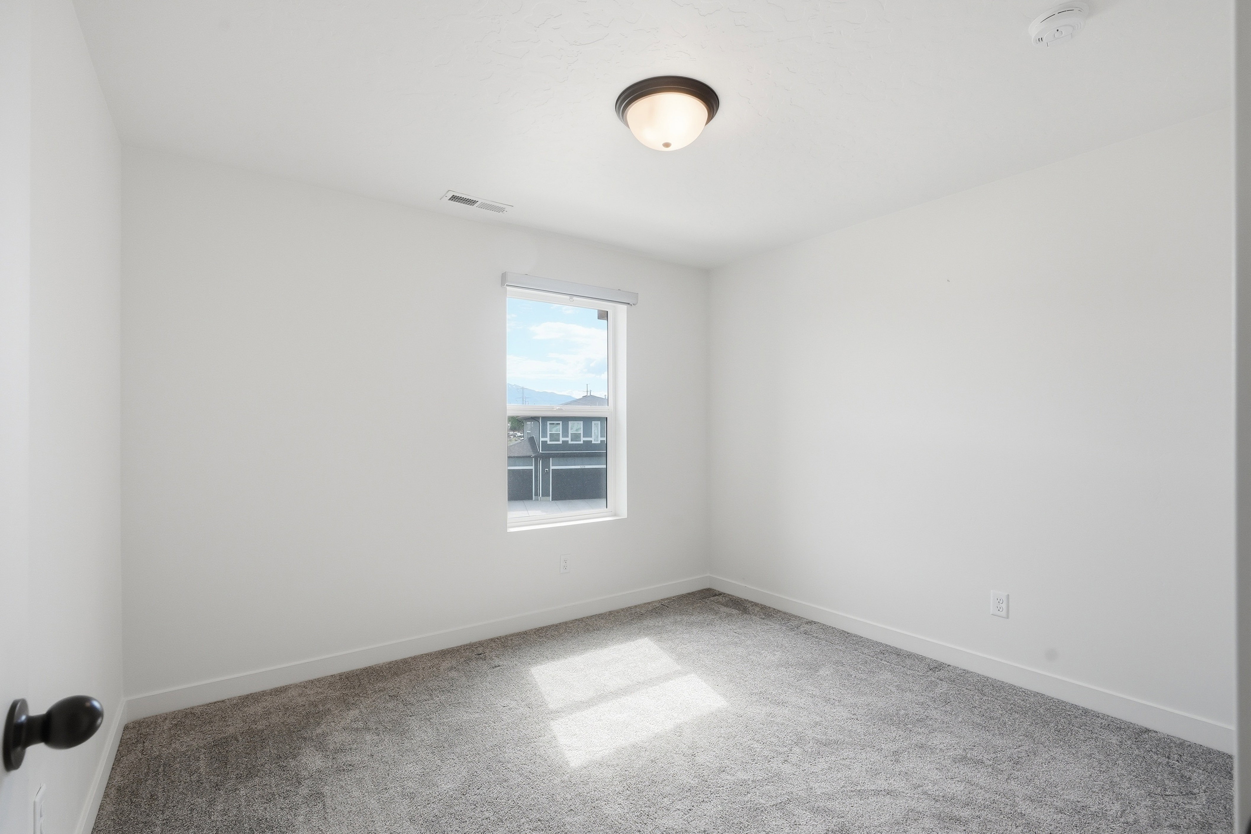 Bright interior space featuring a single window, overhead flush mount lighting, light gray carpeting, crisp white walls, and a visible electrical outlet