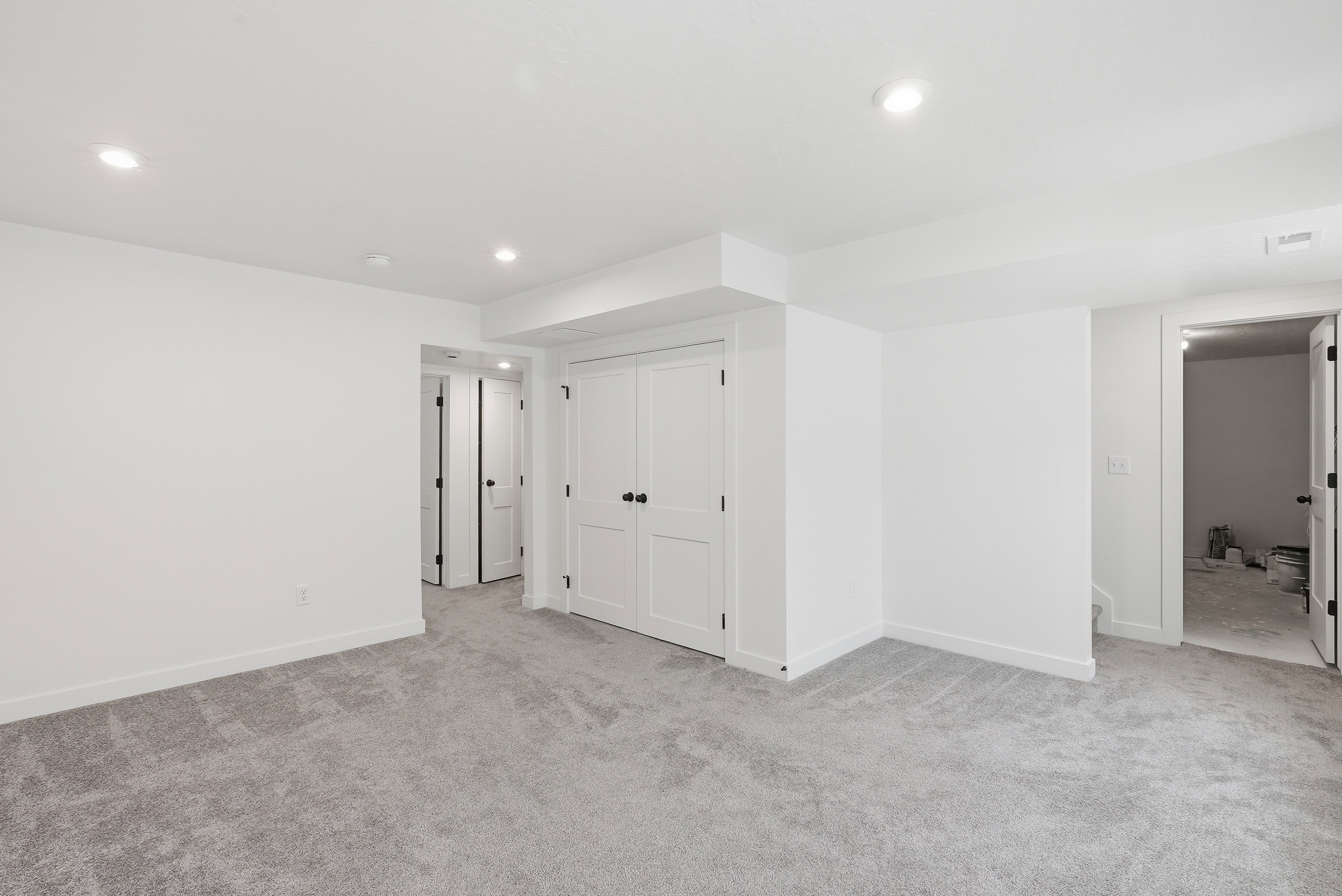 Spacious room featuring light gray carpet flooring, recessed lighting, white walls, and multiple paneled doors with black hardware