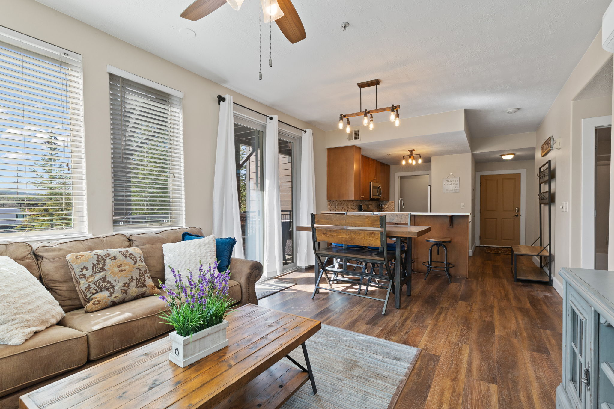 Open-concept living area featuring wood-finish flooring, a ceiling fan with integrated lighting, and multiple windows with blinds
