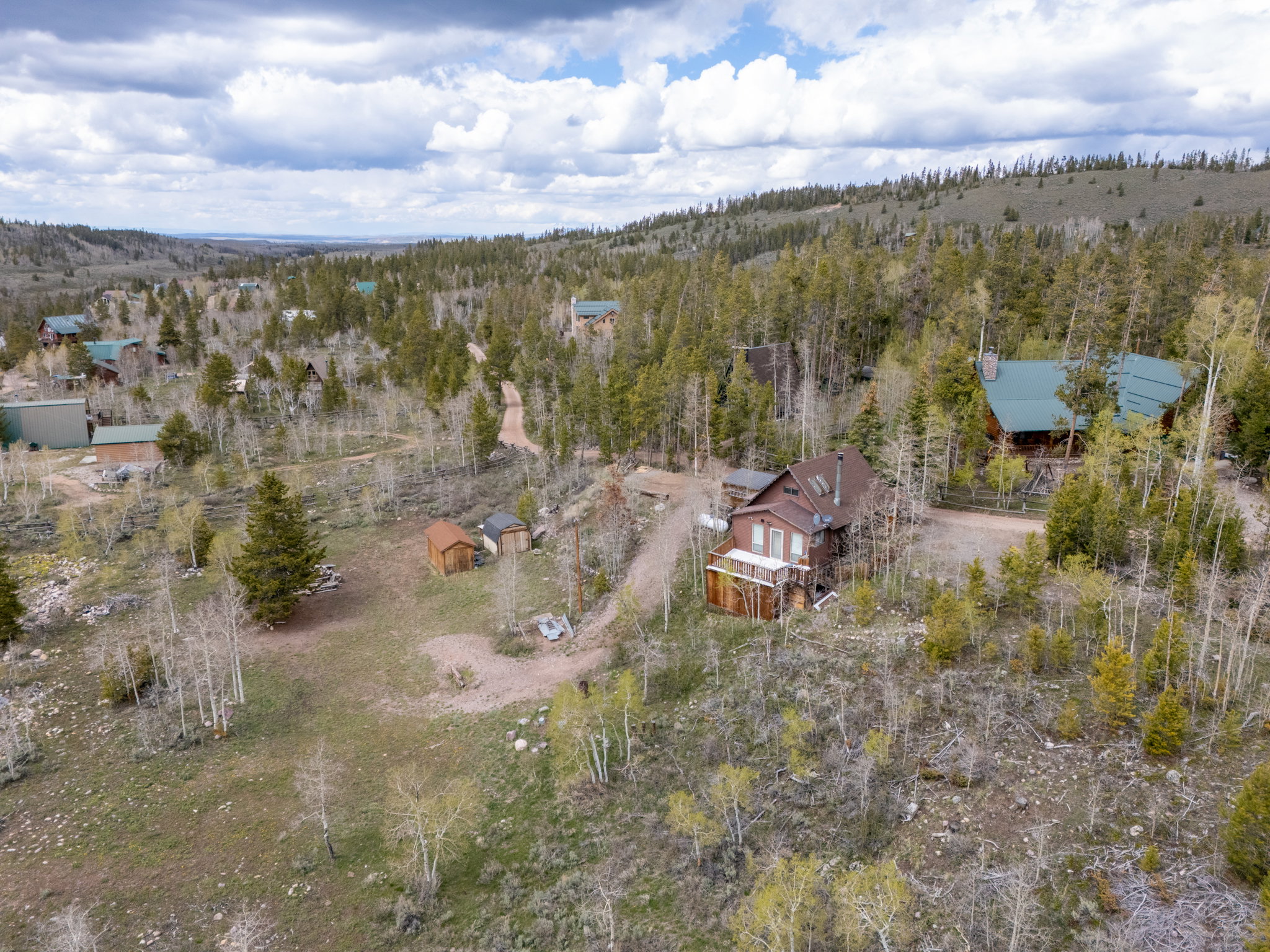Mountain property featuring a brown siding exterior, a prominent green roof, surrounding mature trees, a gravel driveway, and additional outbuildings