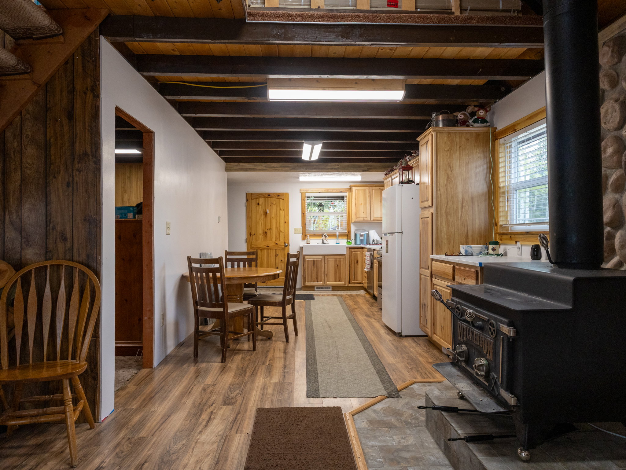 Open-concept kitchen and dining area featuring wood-finish flooring, exposed ceiling beams, and a built-in wood-burning stove