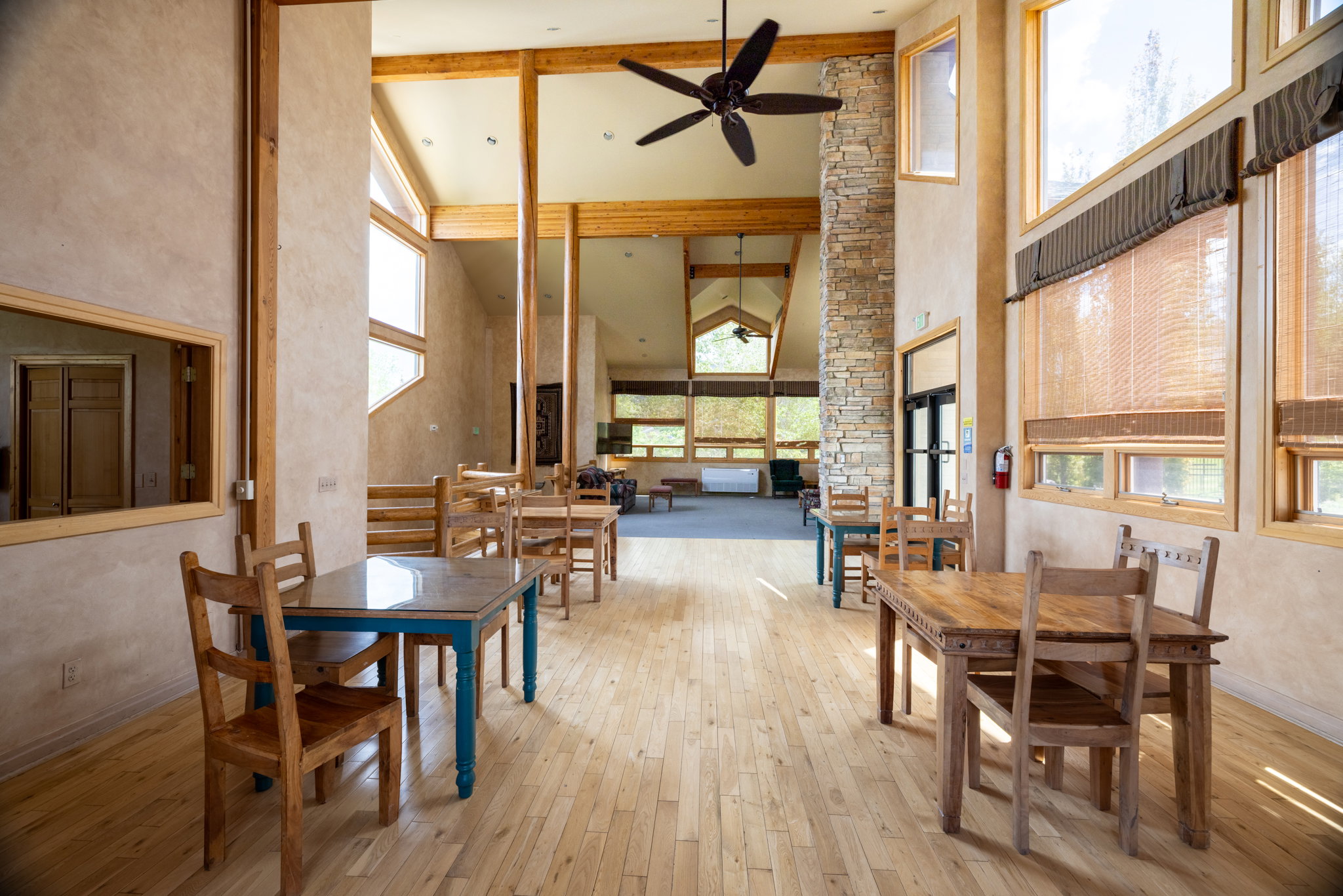 Expansive room featuring light wood-finish flooring, exposed wood beams, a stone accent column, high ceilings, and multiple large windows with natural woven blinds