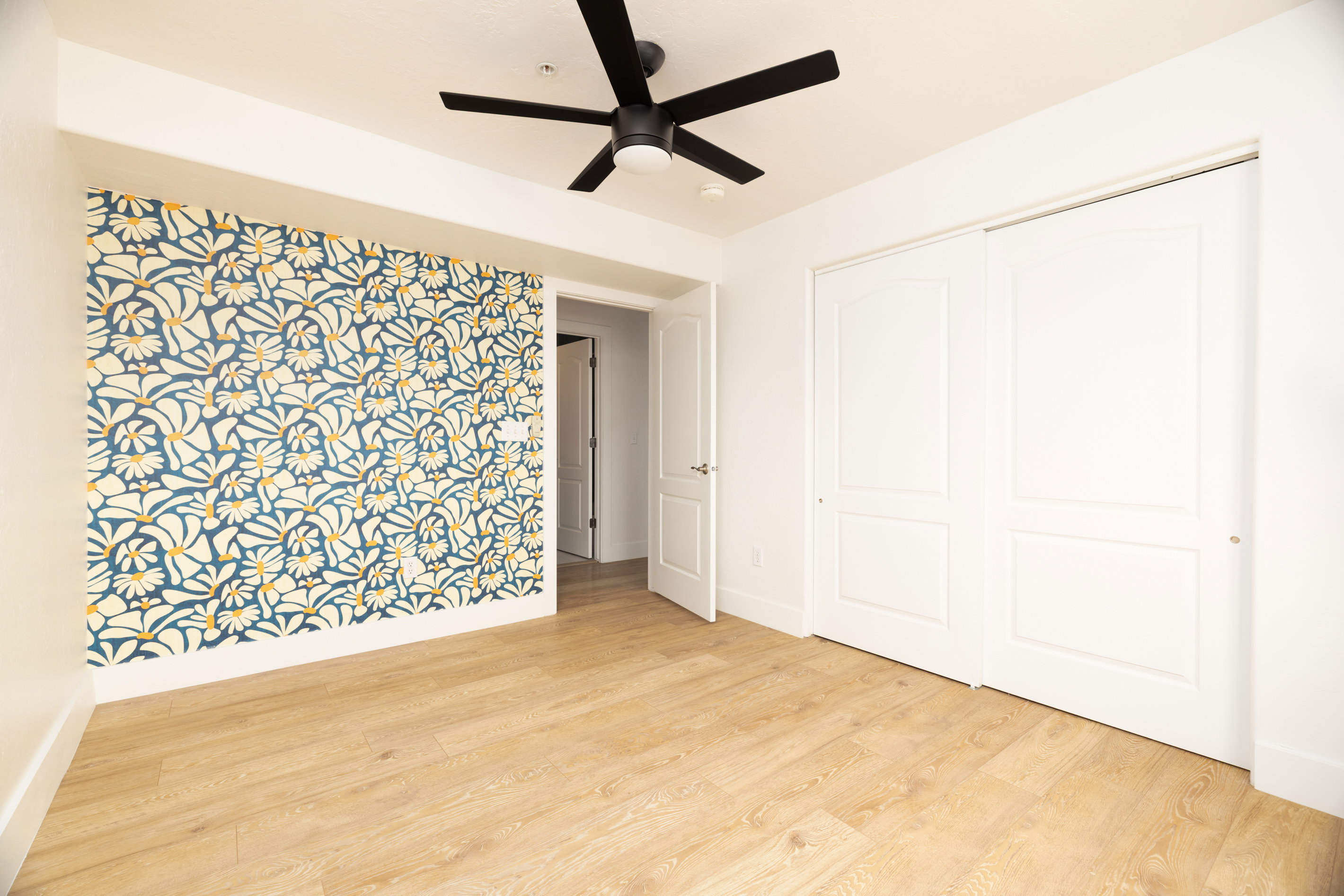 Accent wall featuring a botanical pattern, wood-finish flooring, white sliding closet doors, and a black ceiling fan