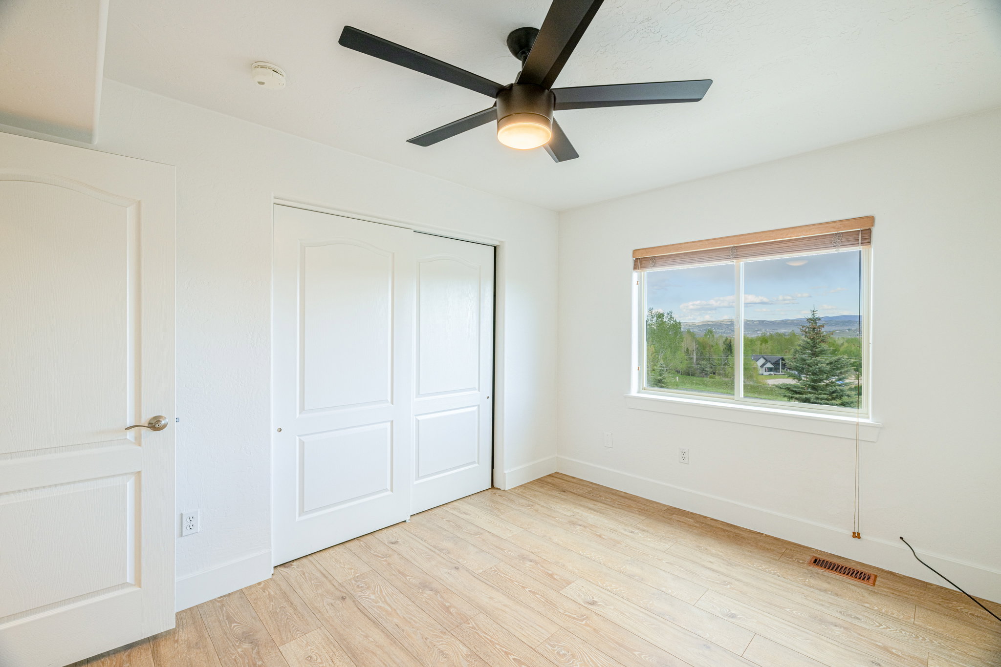 Bright interior space featuring a window with a distant landscape, wood-finish flooring, a contemporary ceiling fan with integrated lighting, a solid-core door with brushed nickel hardware, and bi-fold closet doors