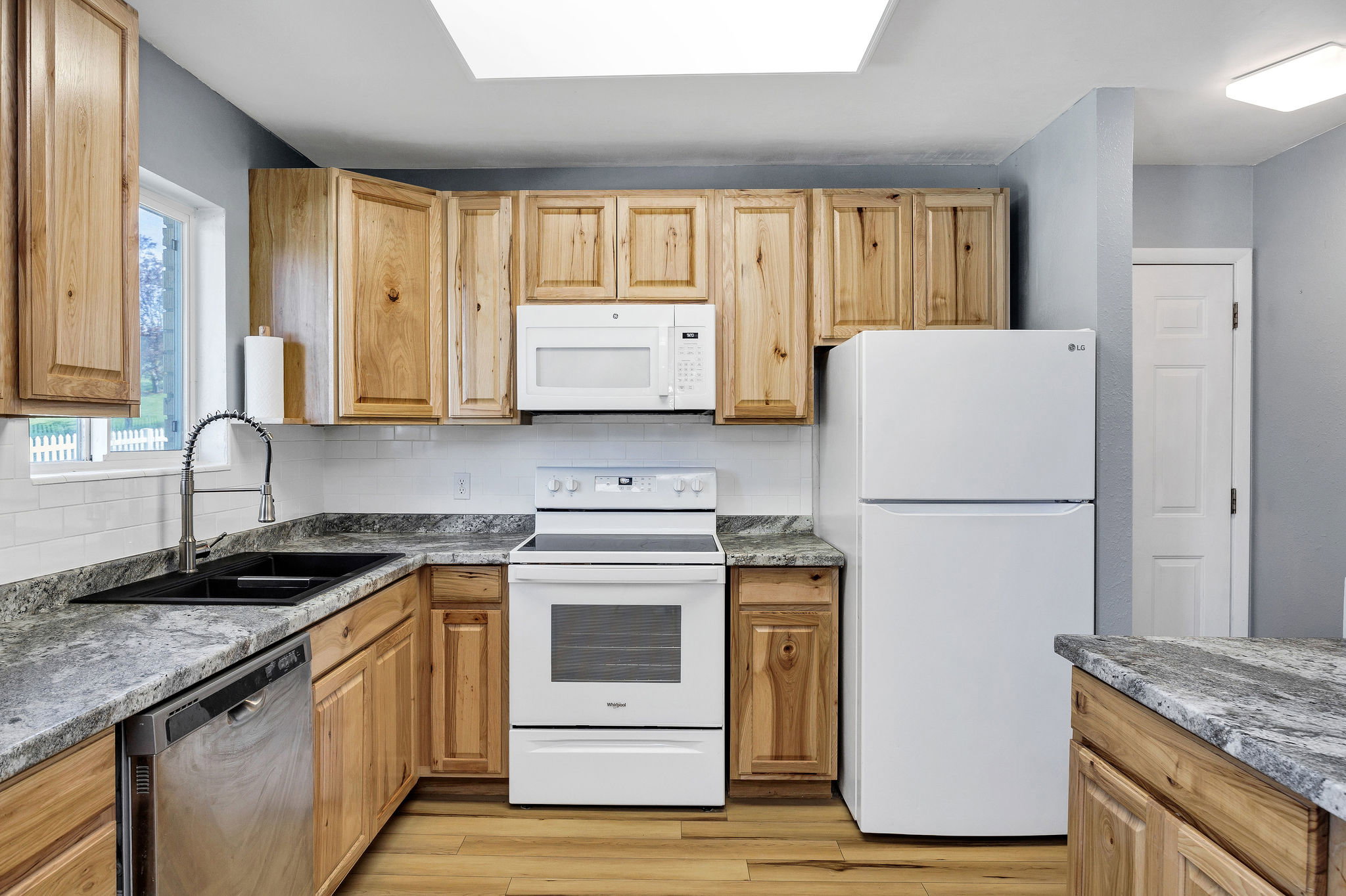 Kitchen featuring natural wood cabinetry, light-toned countertops, white subway tile backsplash, stainless steel dishwasher, and white appliances