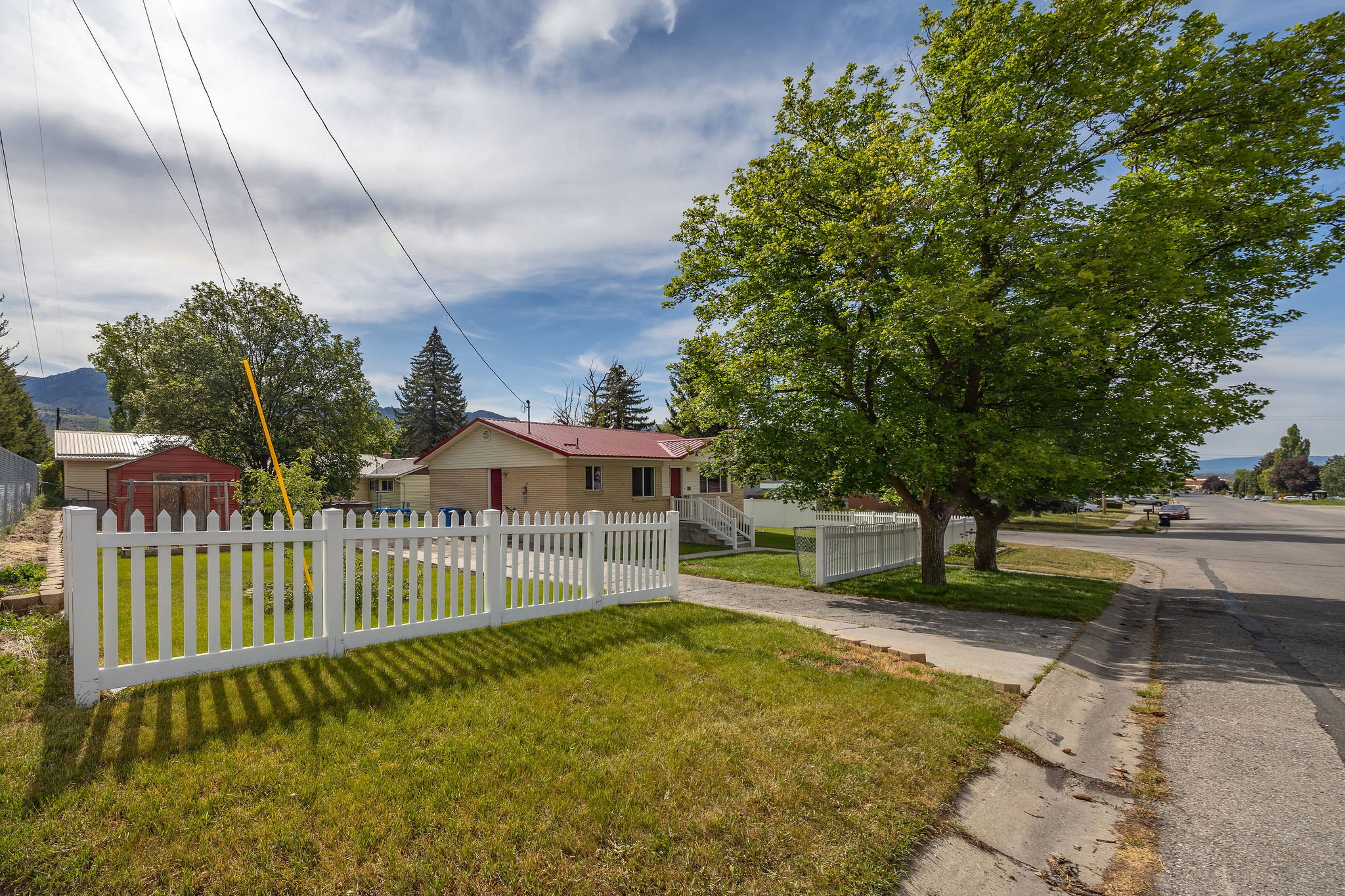 Front yard with white picket fence, mature trees, and a concrete sidewalk