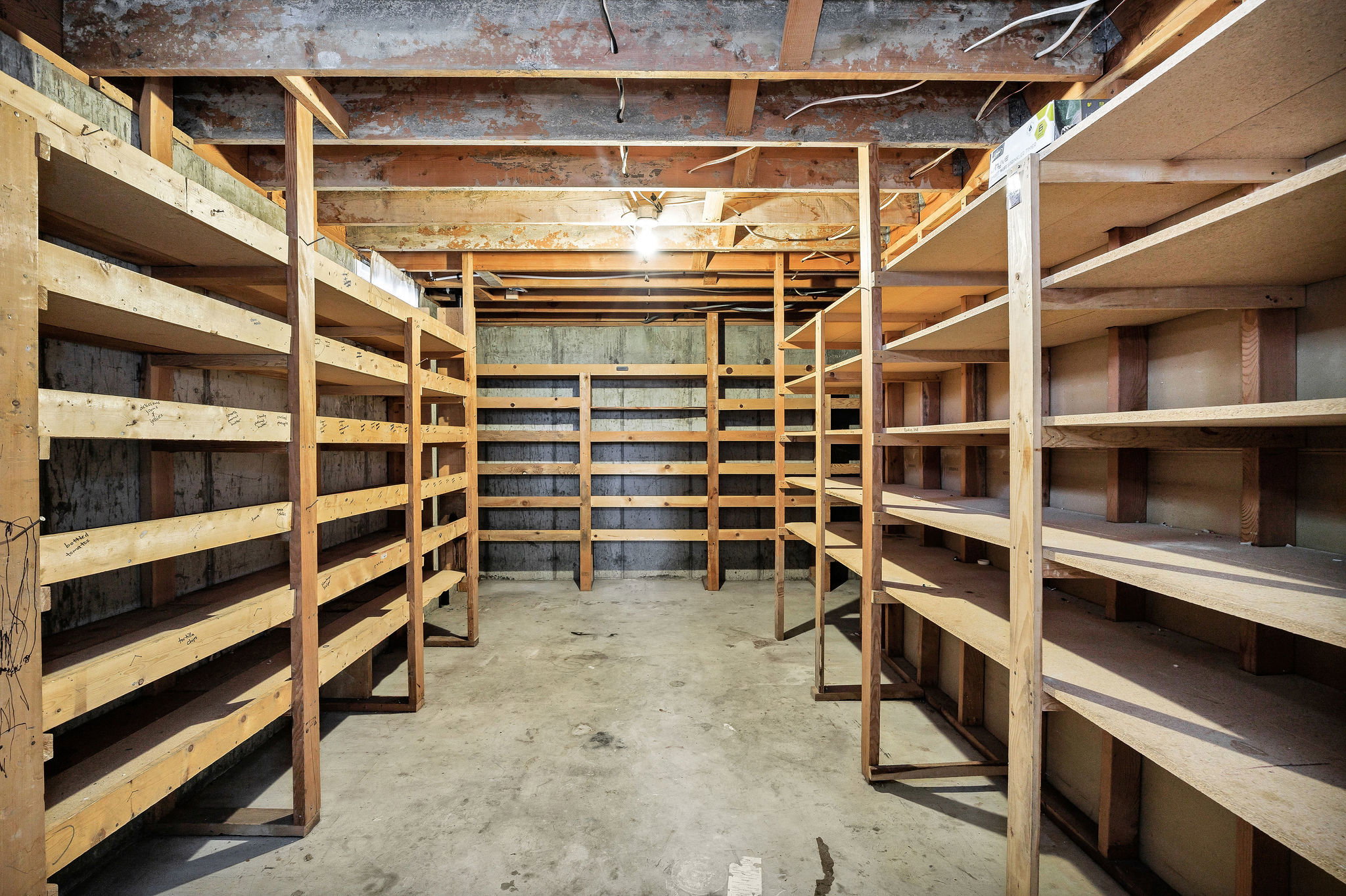 Basement storage area featuring extensive built-in shelving, exposed wood beam ceiling, concrete flooring, and a single ceiling-mounted light fixture