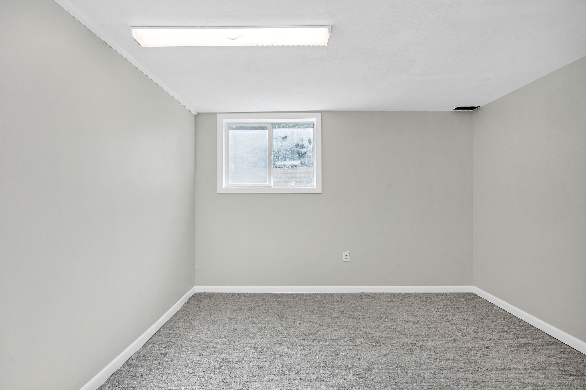 Room featuring gray wall paint, gray carpet flooring, a single window with white trim, white baseboards, and a fluorescent ceiling light fixture