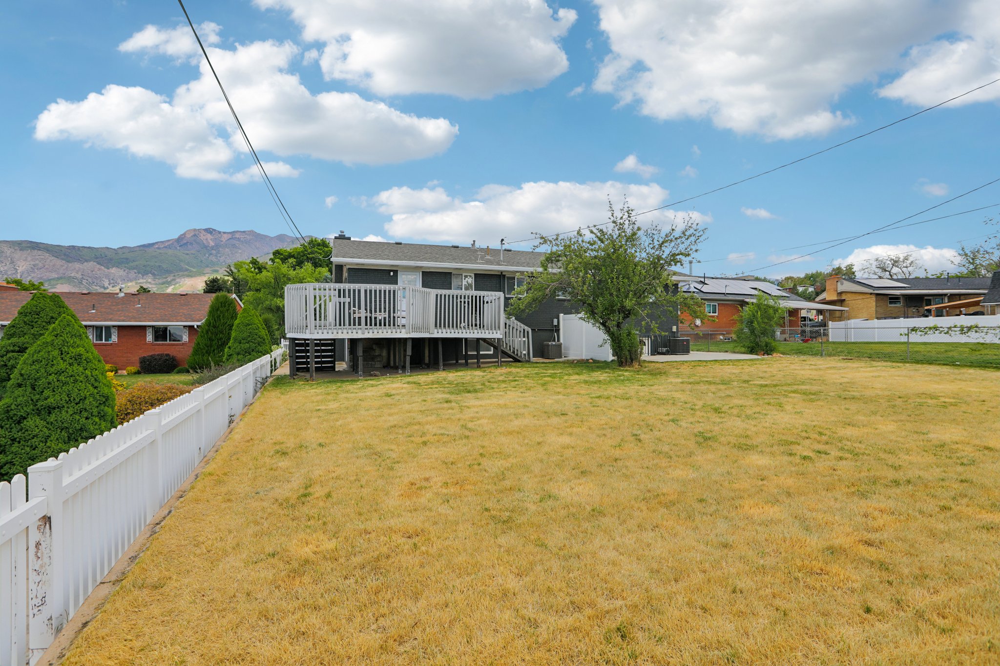 Expansive backyard featuring a white picket fence, a multi-level deck with white railings, a concrete patio, and a mature tree