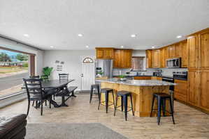 Open-concept living space featuring wood-finish flooring, a kitchen island with seating, natural wood cabinetry, stainless steel appliances, and a large picture window