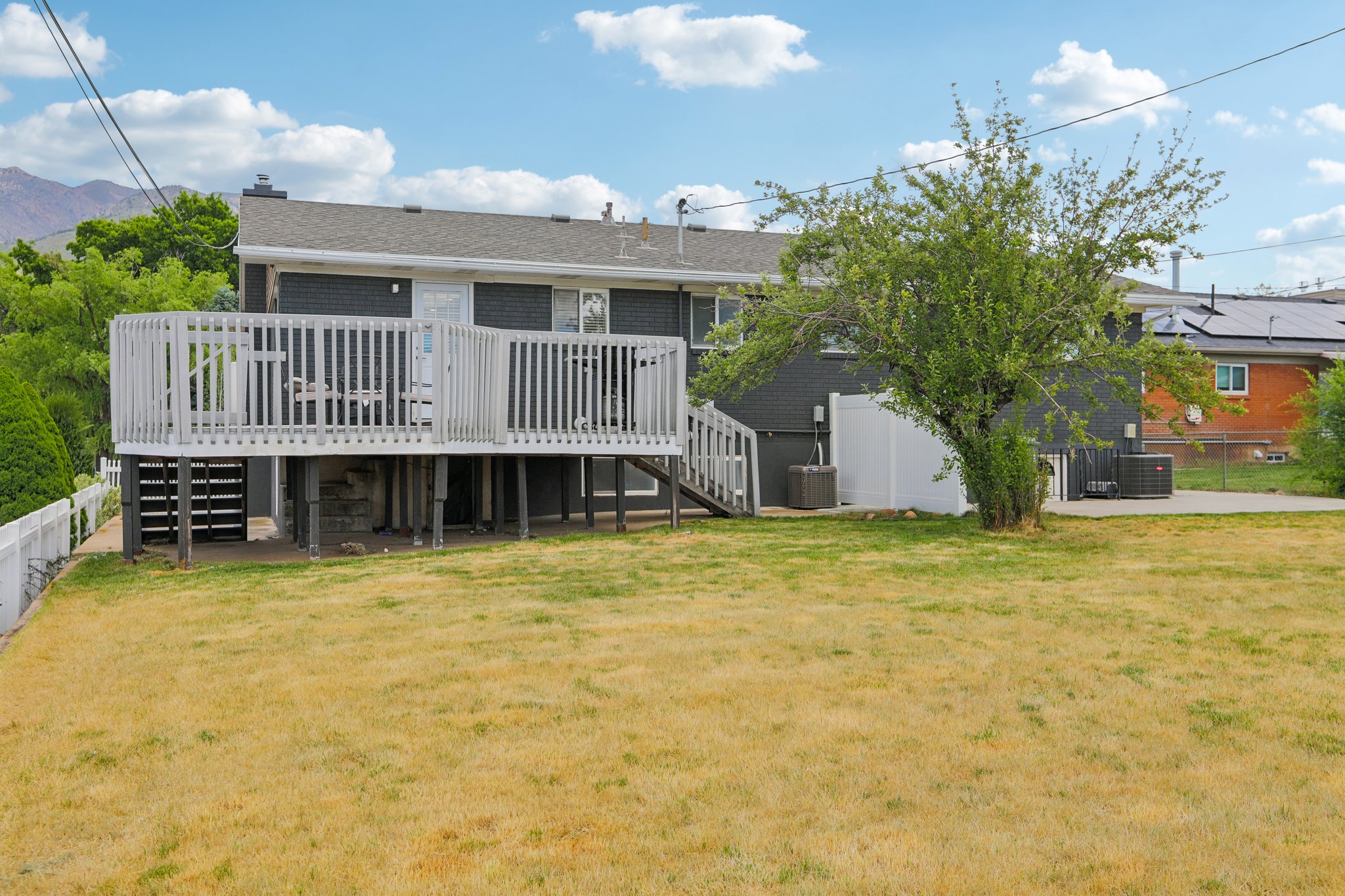 Rear exterior showcasing a spacious elevated deck with white railing, a mature shade tree, a concrete patio, and a white privacy fence