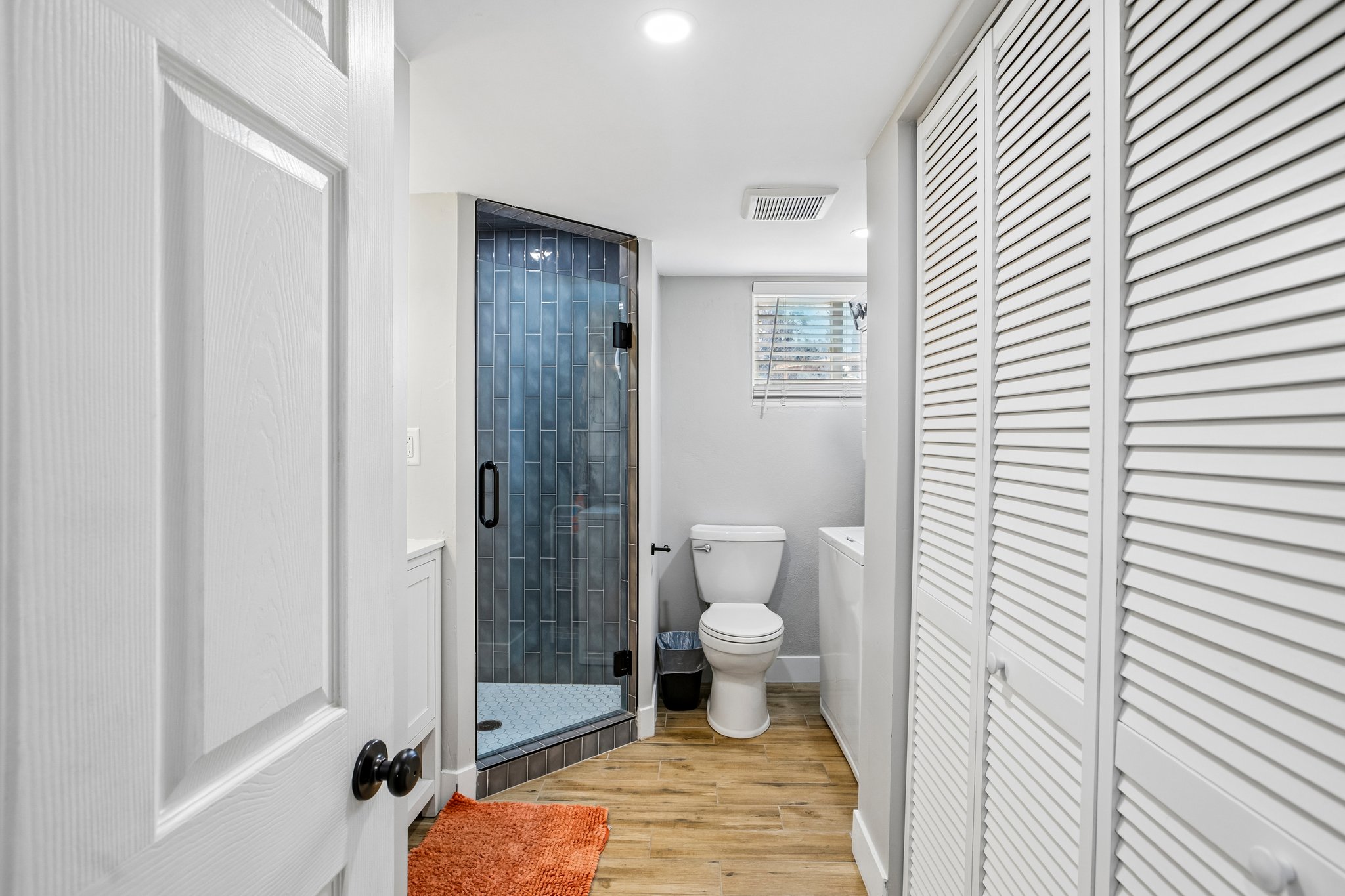 Bathroom featuring a glass-enclosed shower with dark vertical tile, wood-finish flooring, a white toilet, and louvered closet doors