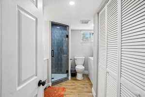 Bathroom featuring a glass-enclosed shower with dark vertical tile, wood-finish flooring, a white toilet, and louvered closet doors