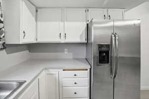 White cabinetry with black hardware, light-toned countertops, stainless steel side-by-side refrigerator with exterior dispenser, integrated stainless steel sink, and drawer storage