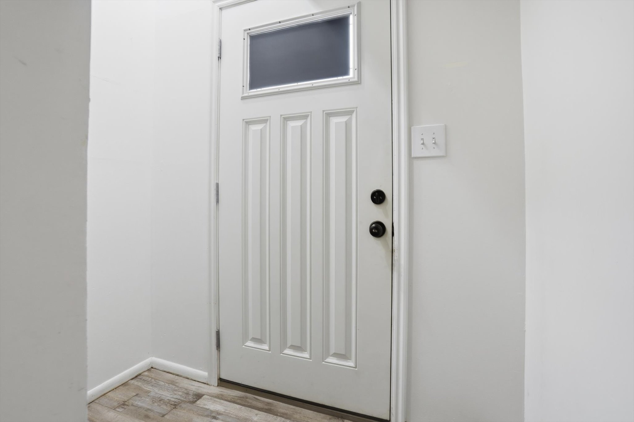 White paneled door with an upper window insert, featuring black hardware and wood-finish flooring