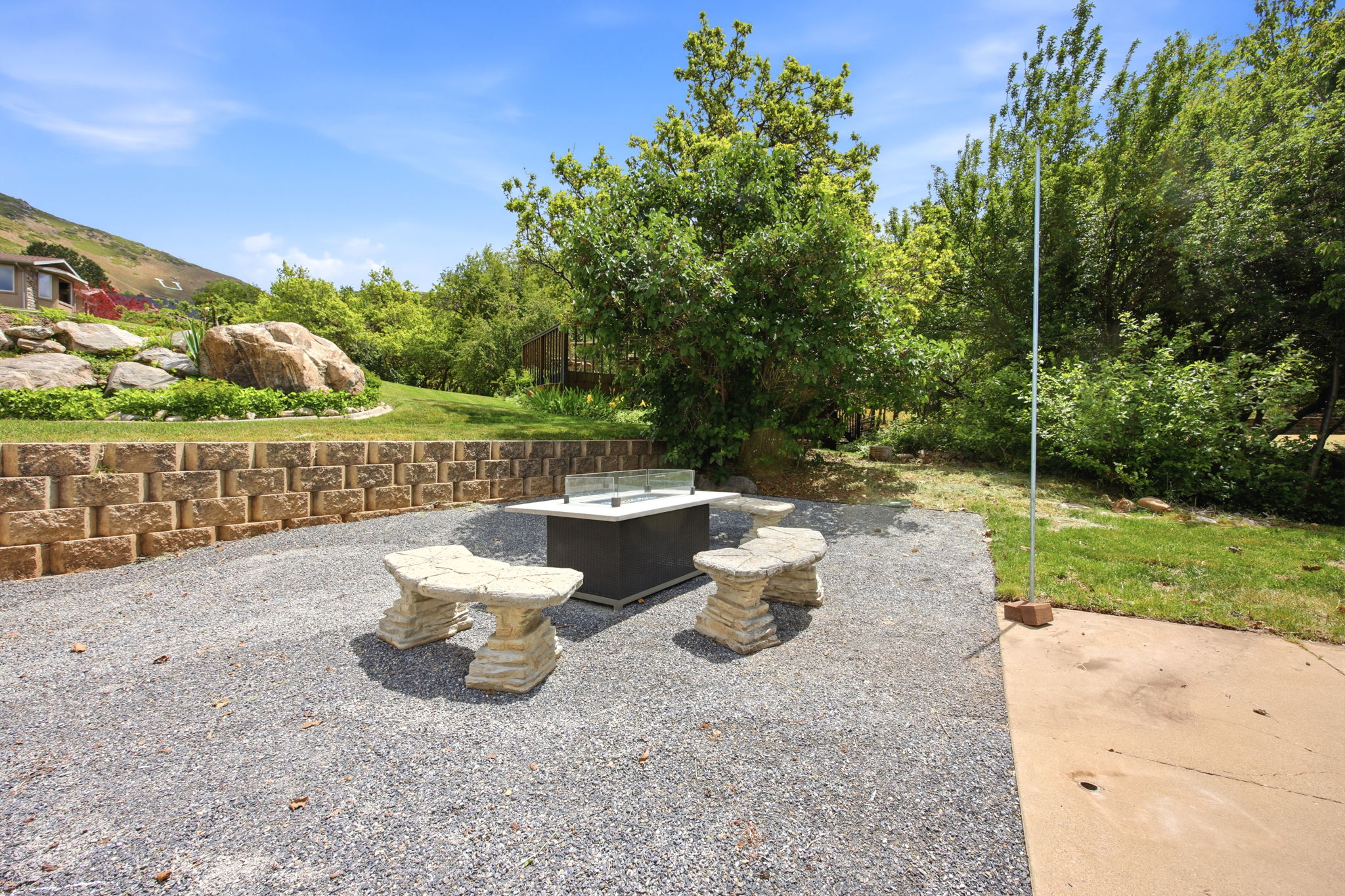 Outdoor entertainment area featuring a rectangular fire pit with glass surround, two stone benches, and a gravel patio surface