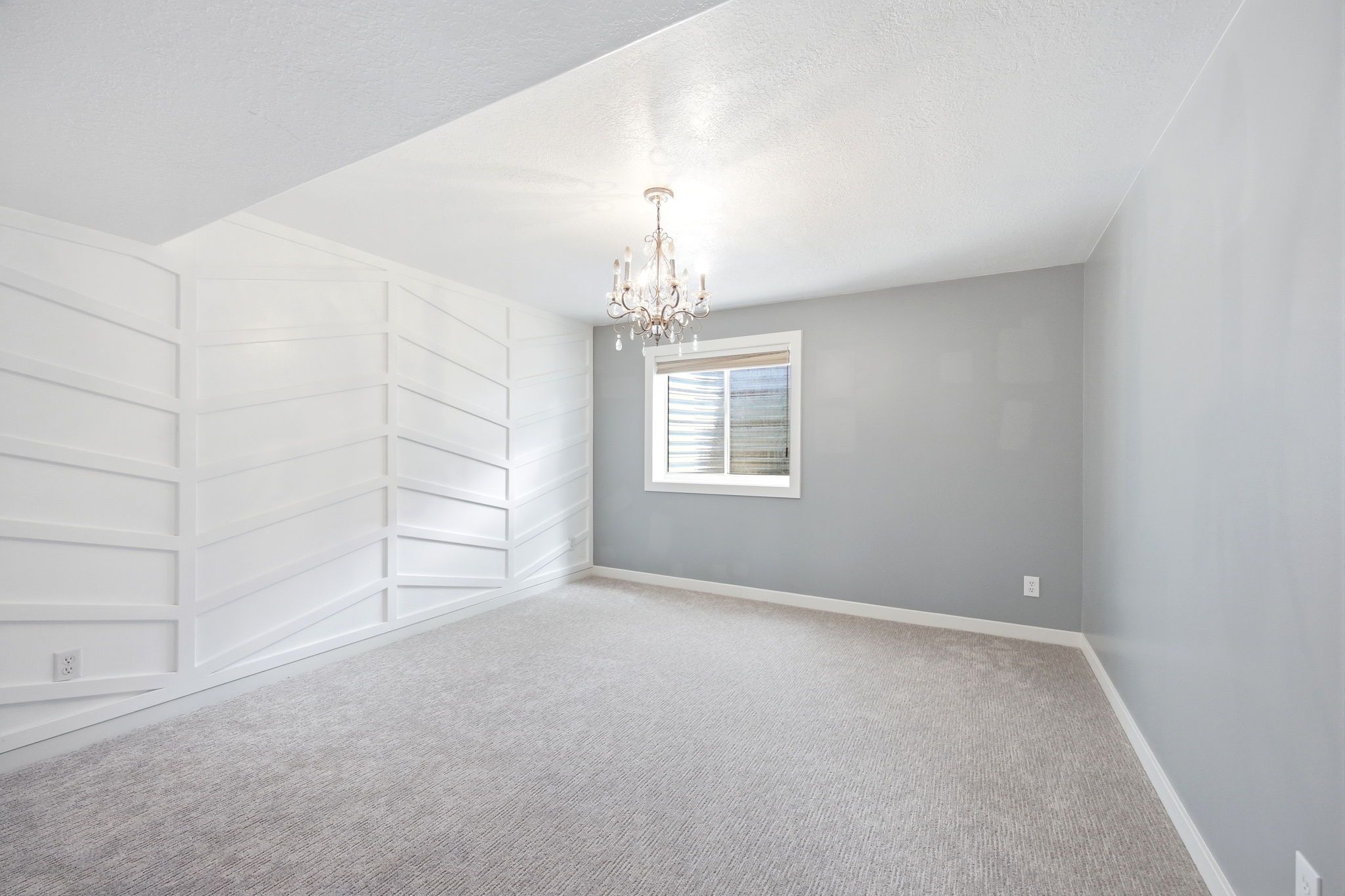 Spacious room featuring a decorative accent wall with geometric paneling, light gray carpeting, and a window with horizontal blinds