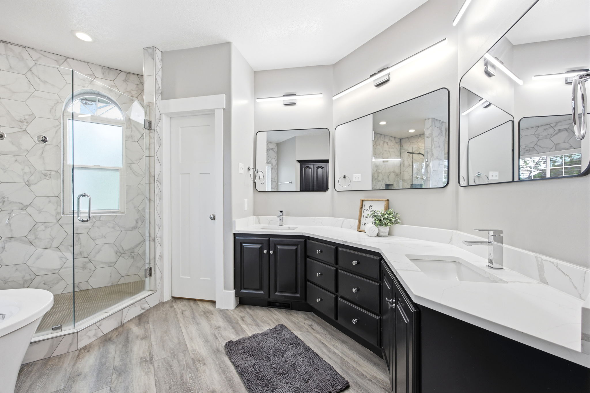 Contemporary bathroom featuring a spacious glass-enclosed shower with hexagon tile surround and arched window, wood-finish flooring, dual vanity with black cabinetry, white countertops, and sleek wall-mounted mirrors with integrated lighting