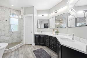 Contemporary bathroom featuring a spacious glass-enclosed shower with hexagon tile surround and arched window, wood-finish flooring, dual vanity with black cabinetry, white countertops, and sleek wall-mounted mirrors with integrated lighting