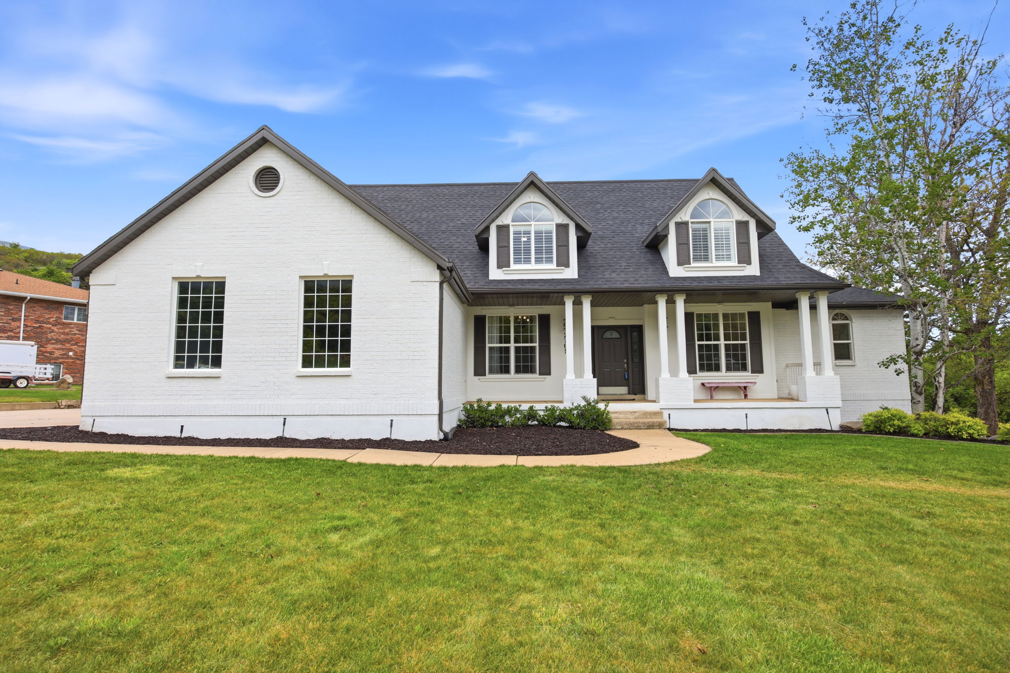 White brick facade featuring dormer windows, dark roof, dark shutters, covered front porch with columns, and a landscaped front yard
