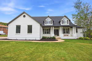 White brick facade featuring dormer windows, dark roof, dark shutters, covered front porch with columns, and a landscaped front yard