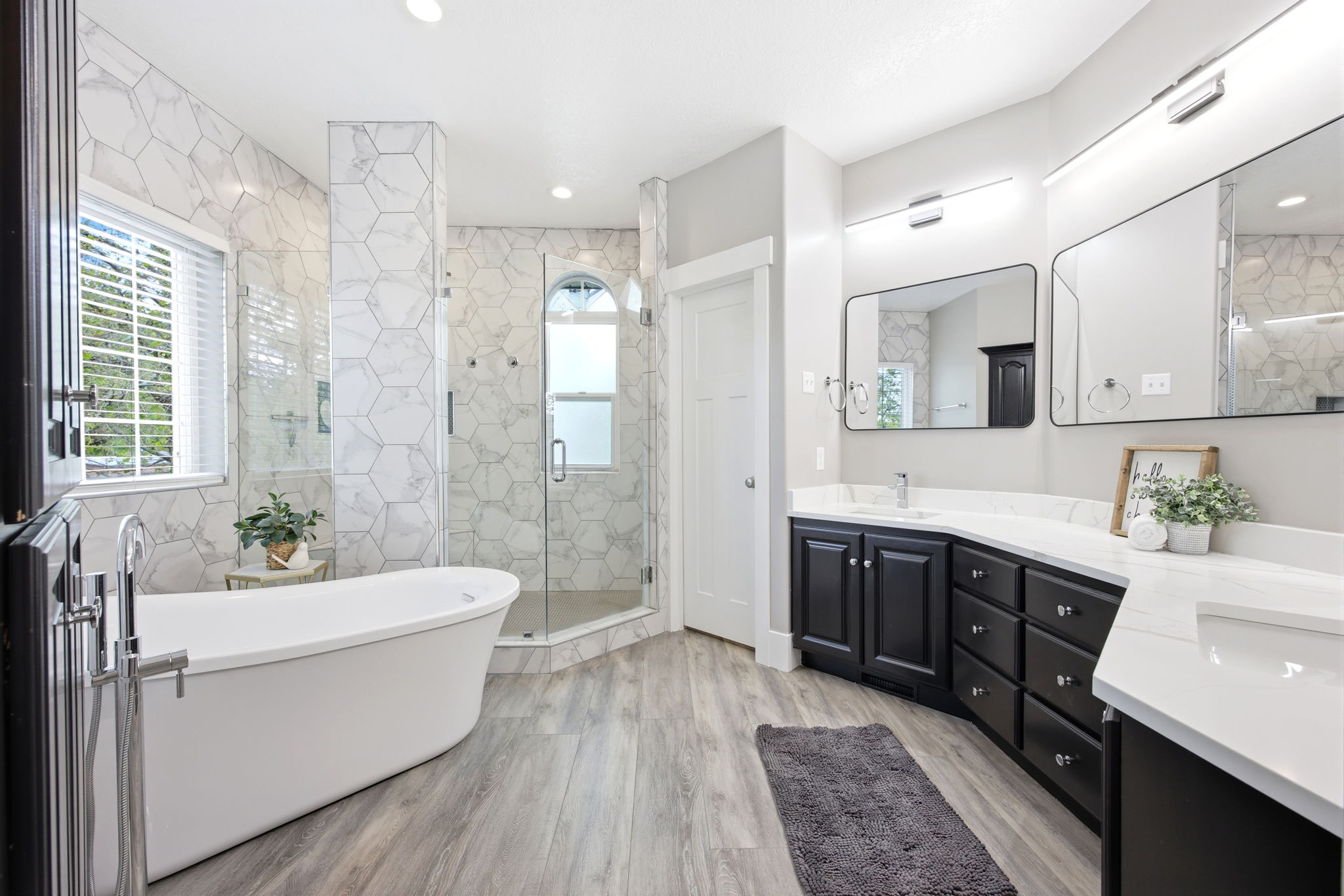 Spacious bathroom featuring a freestanding tub, glass-enclosed shower, dual vanities with dark cabinetry, and light wood-finish flooring