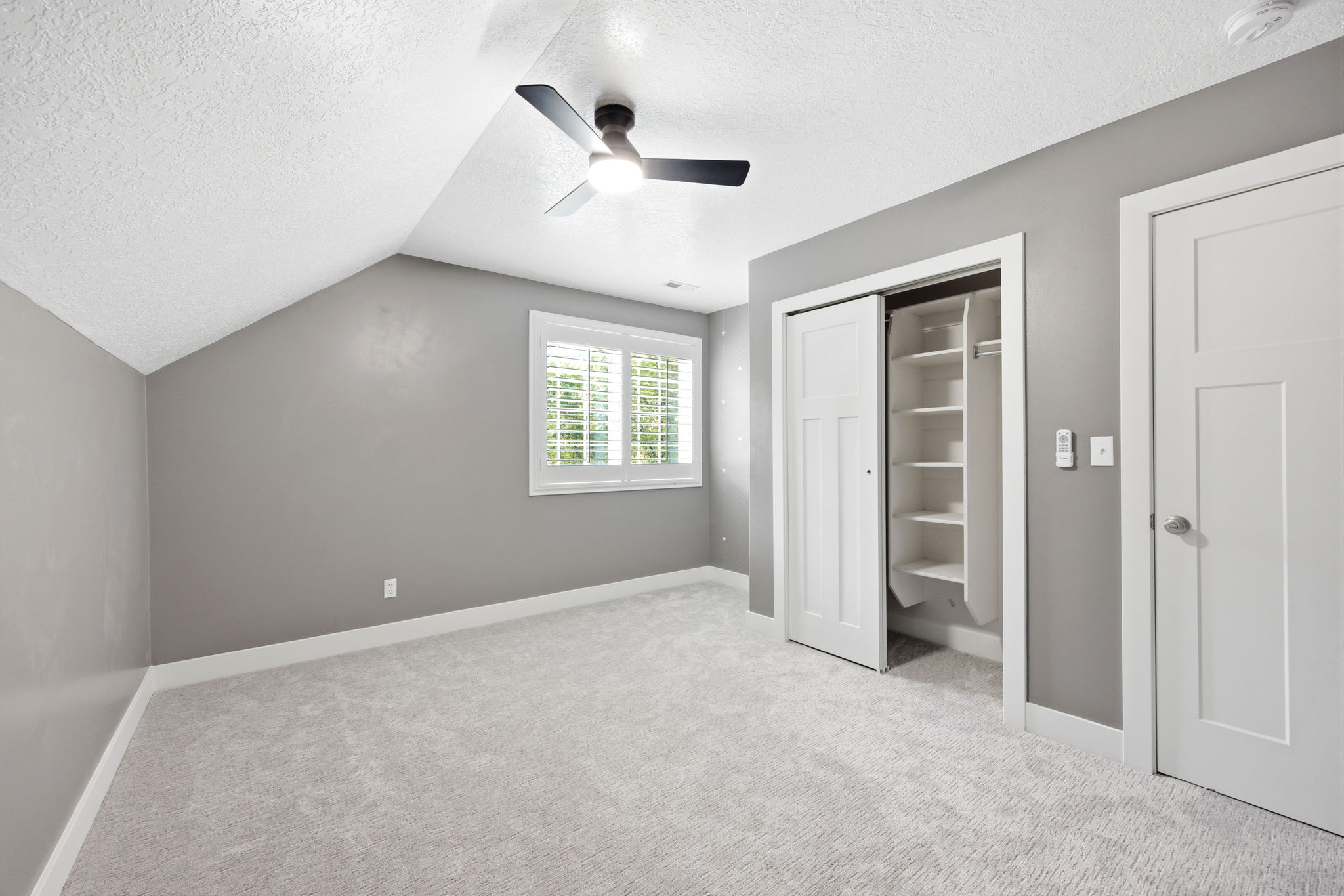 Carpeted room featuring a ceiling fan with integrated lighting, a window with white plantation shutters, and a closet with shelving and a hanging rod