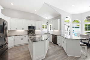 Bright kitchen featuring white cabinetry, black stone countertops, a central island, stainless steel appliances, and wood-finish flooring