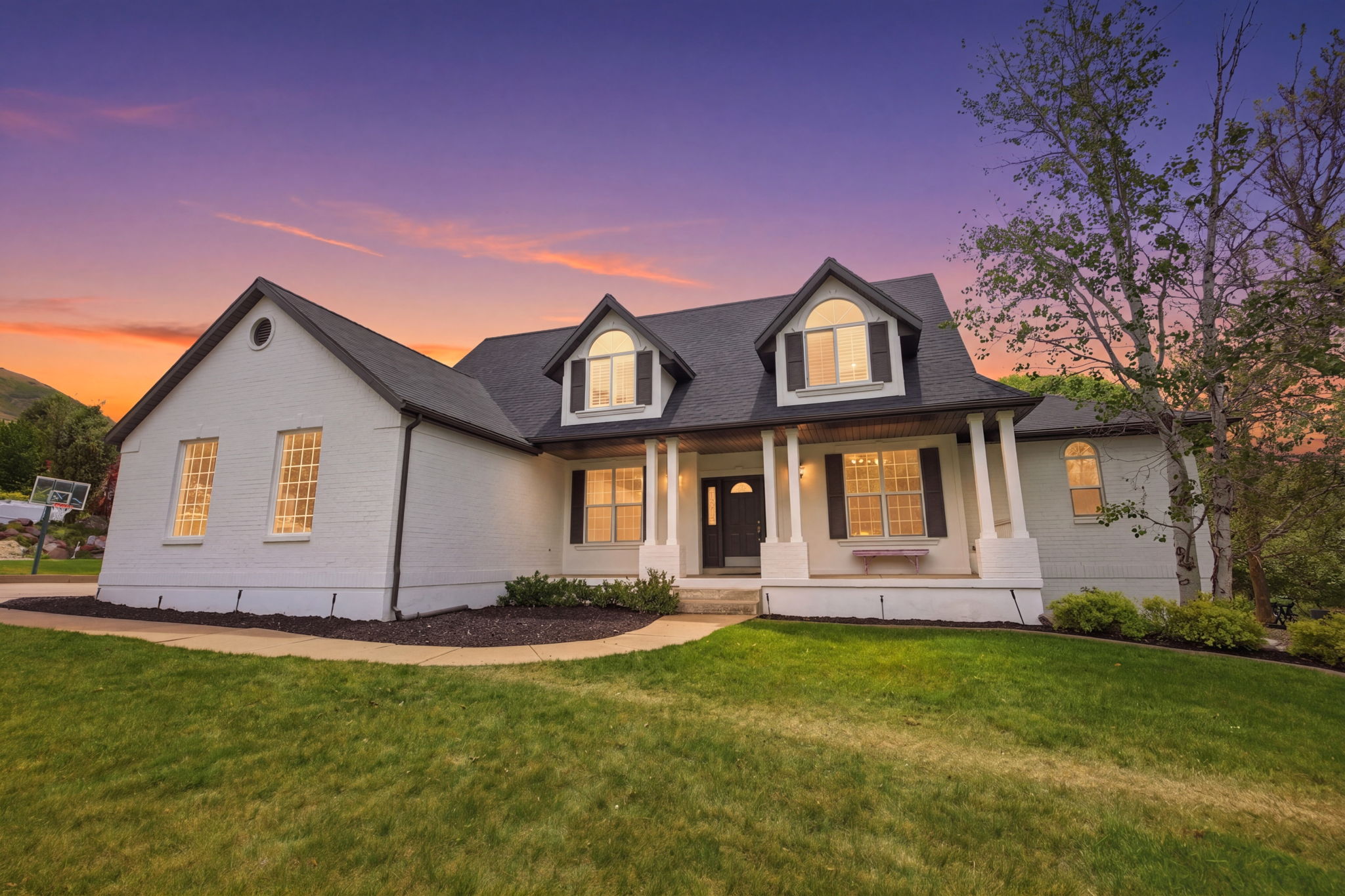 Contemporary residence featuring a white brick exterior, dark pitched roof, gabled dormers with arched windows, a covered front porch with white columns, and a landscaped front yard with a manicured lawn