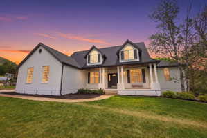 Contemporary residence featuring a white brick exterior, dark pitched roof, gabled dormers with arched windows, a covered front porch with white columns, and a landscaped front yard with a manicured lawn