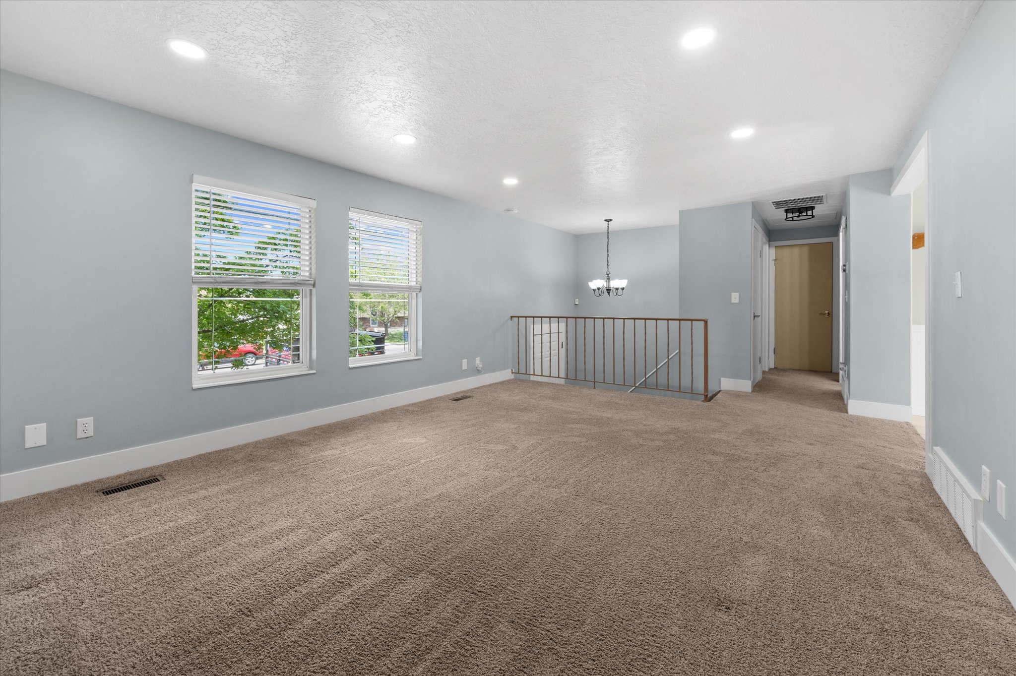 Spacious room with plush carpeting, two large windows with blinds, recessed lighting, and a partial baluster railing