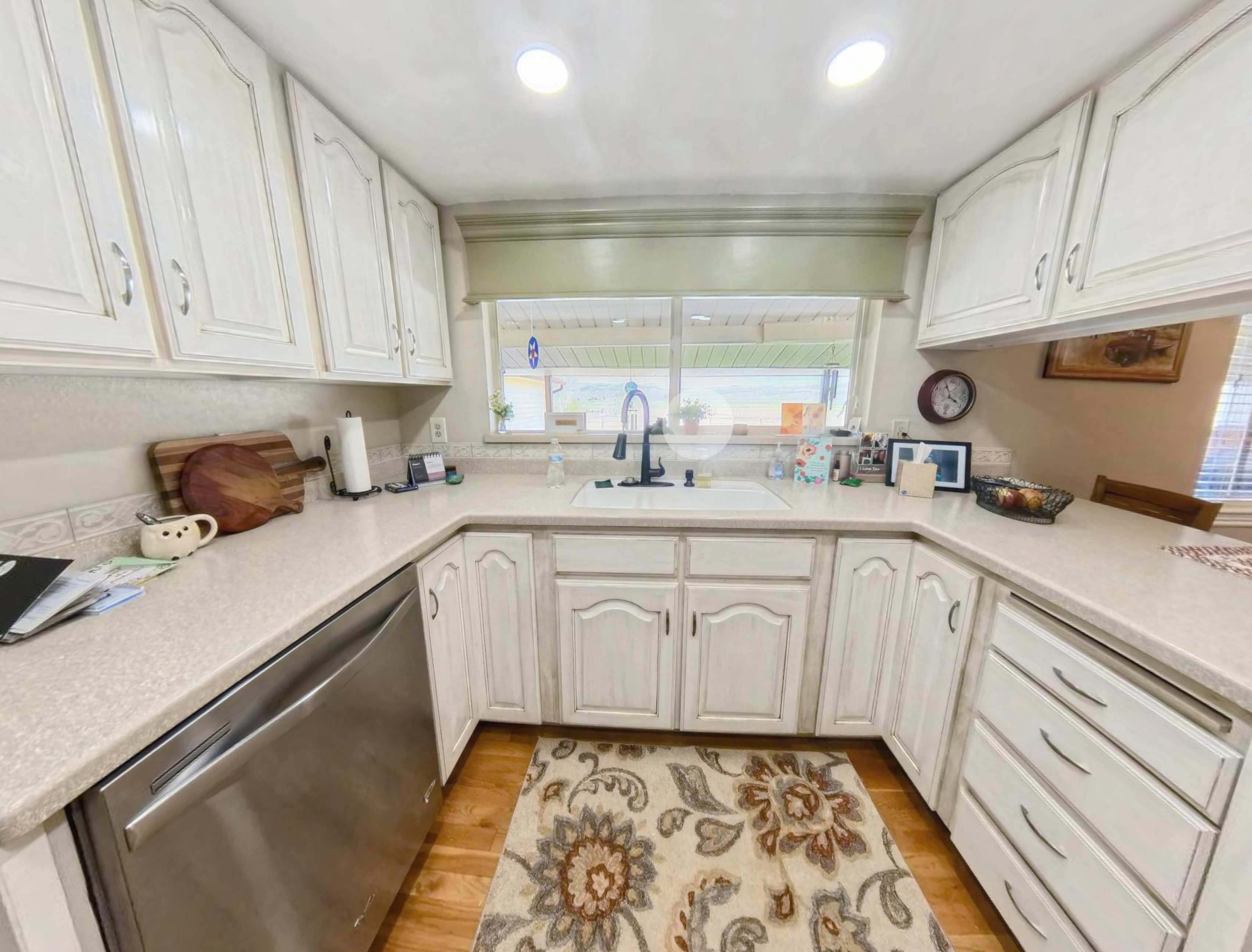 U-shaped kitchen featuring wood-finish flooring, white cabinetry with arched panels, light-toned countertops, and a stainless steel dishwasher